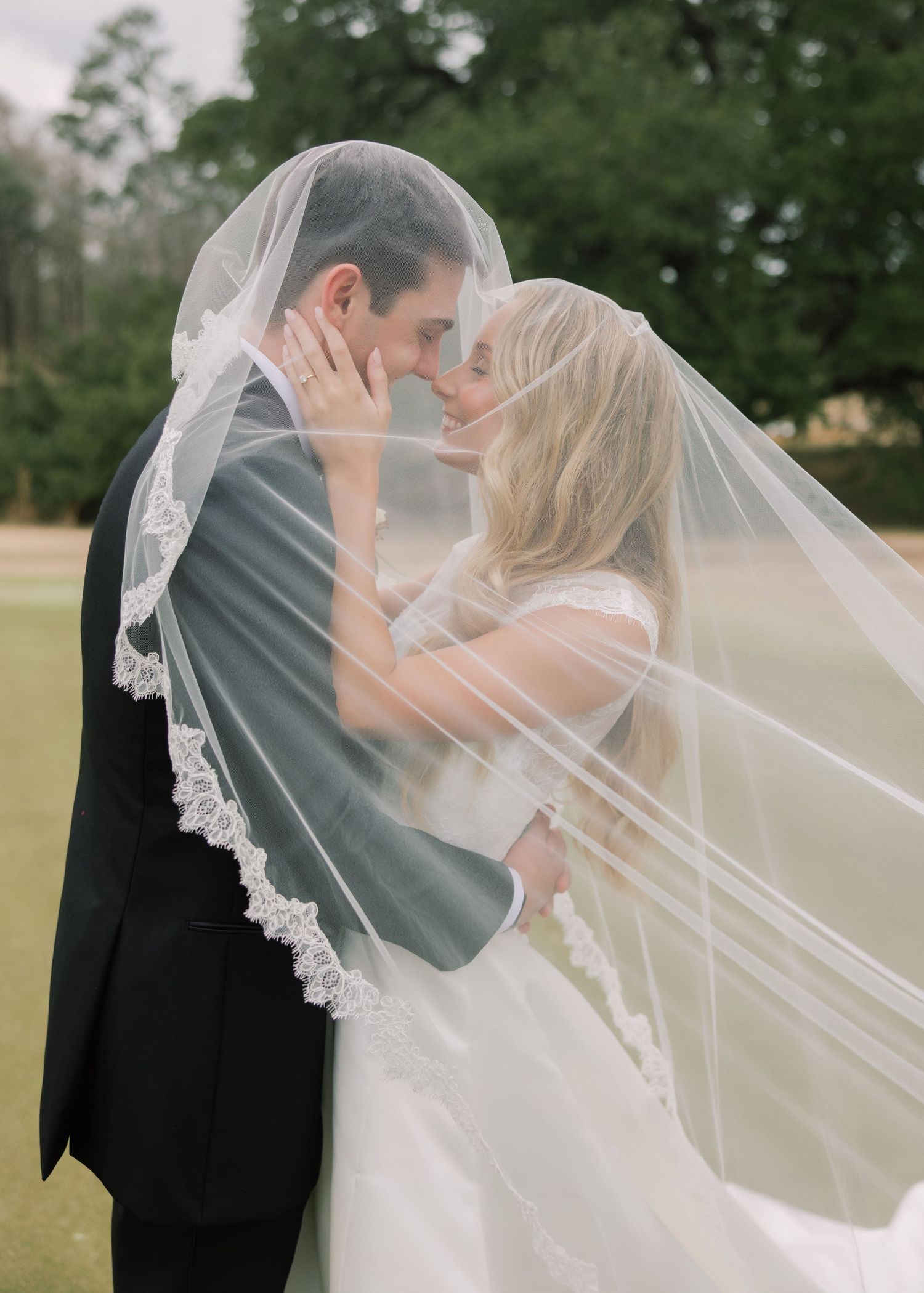 A romantic wedding couple shares a tender kiss under a flowing lace veil in an outdoor garden setting.