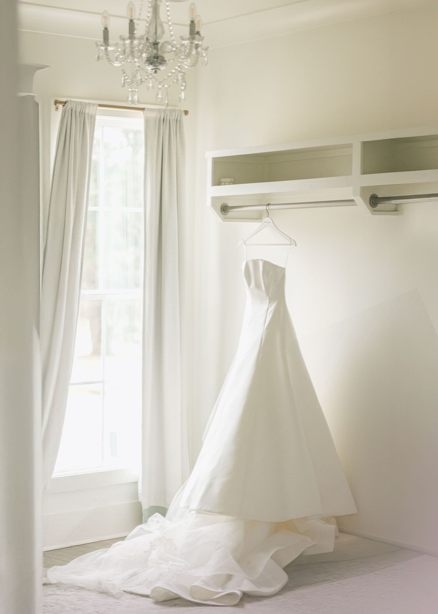A row of elegant white wedding dresses hanging in a bright room with crystal chandeliers and white curtains.