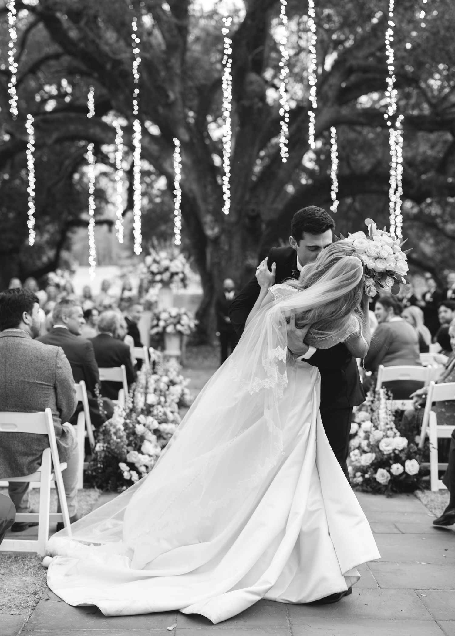 A romantic black and white wedding photo showing a couple's first dance under twinkling lights and trees at an outdoor ceremony.