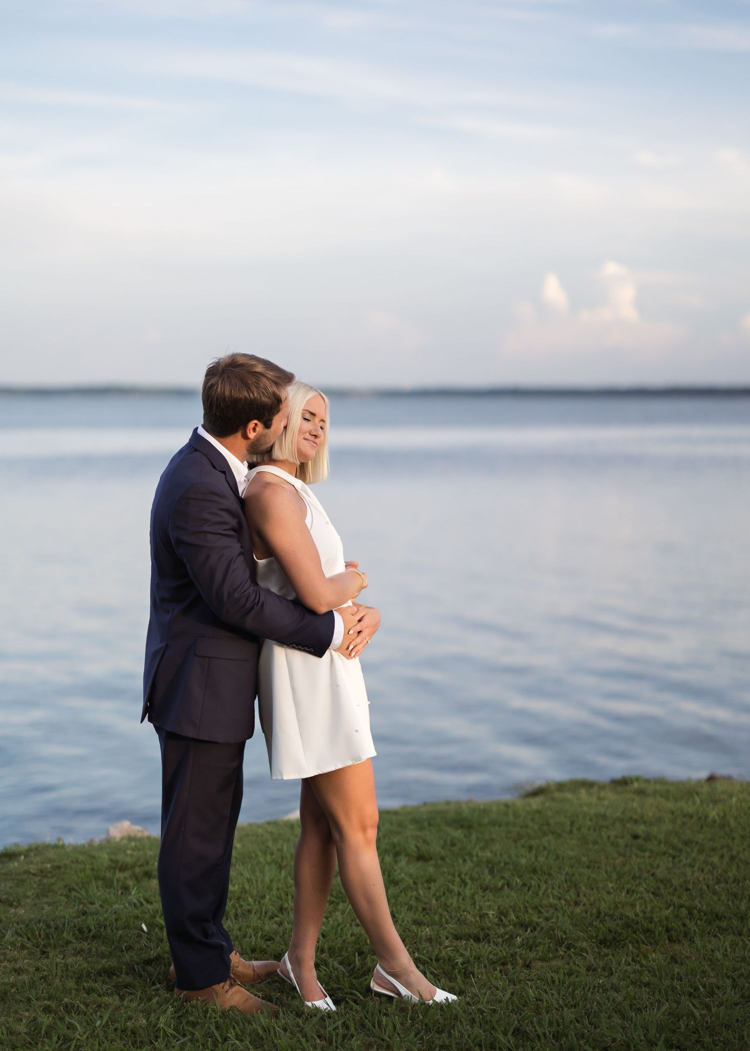 Couple in formal wear sharing romantic moments on grassy waterfront.