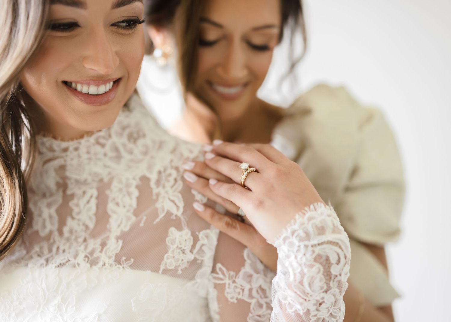 A bride in an elegant lace wedding dress smiles while getting ready with her bridesmaids in matching robes.