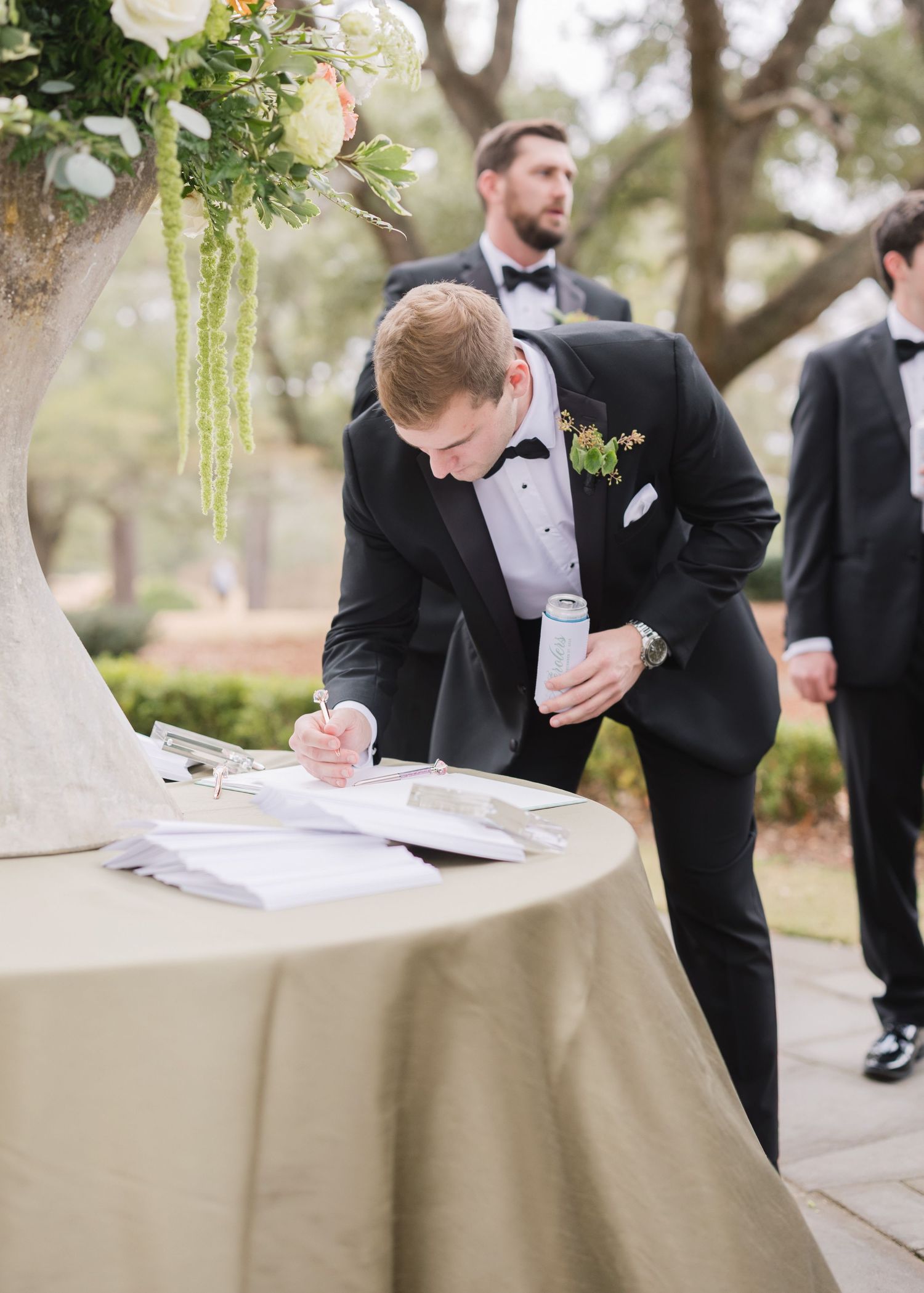 Groomsmen in black tuxedos sign wedding documents at an outdoor ceremony table covered with a beige tablecloth.