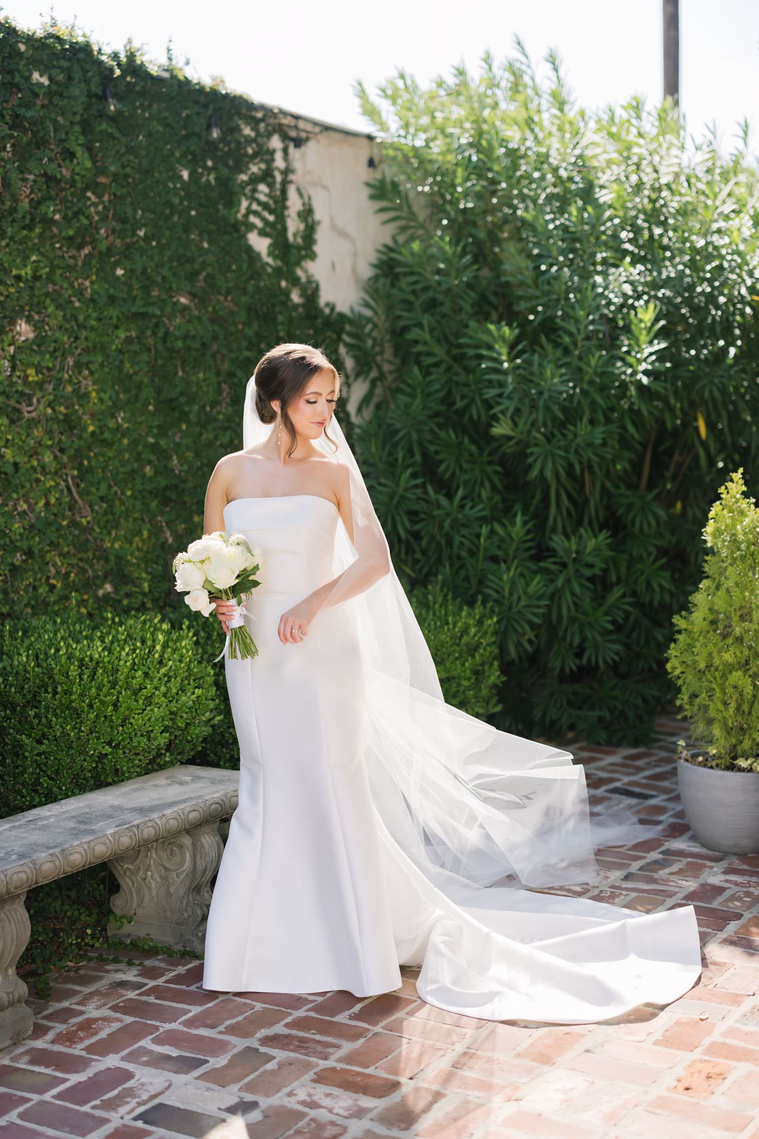 A bride in a white wedding dress and veil poses gracefully in a garden setting with brick paving and greenery.