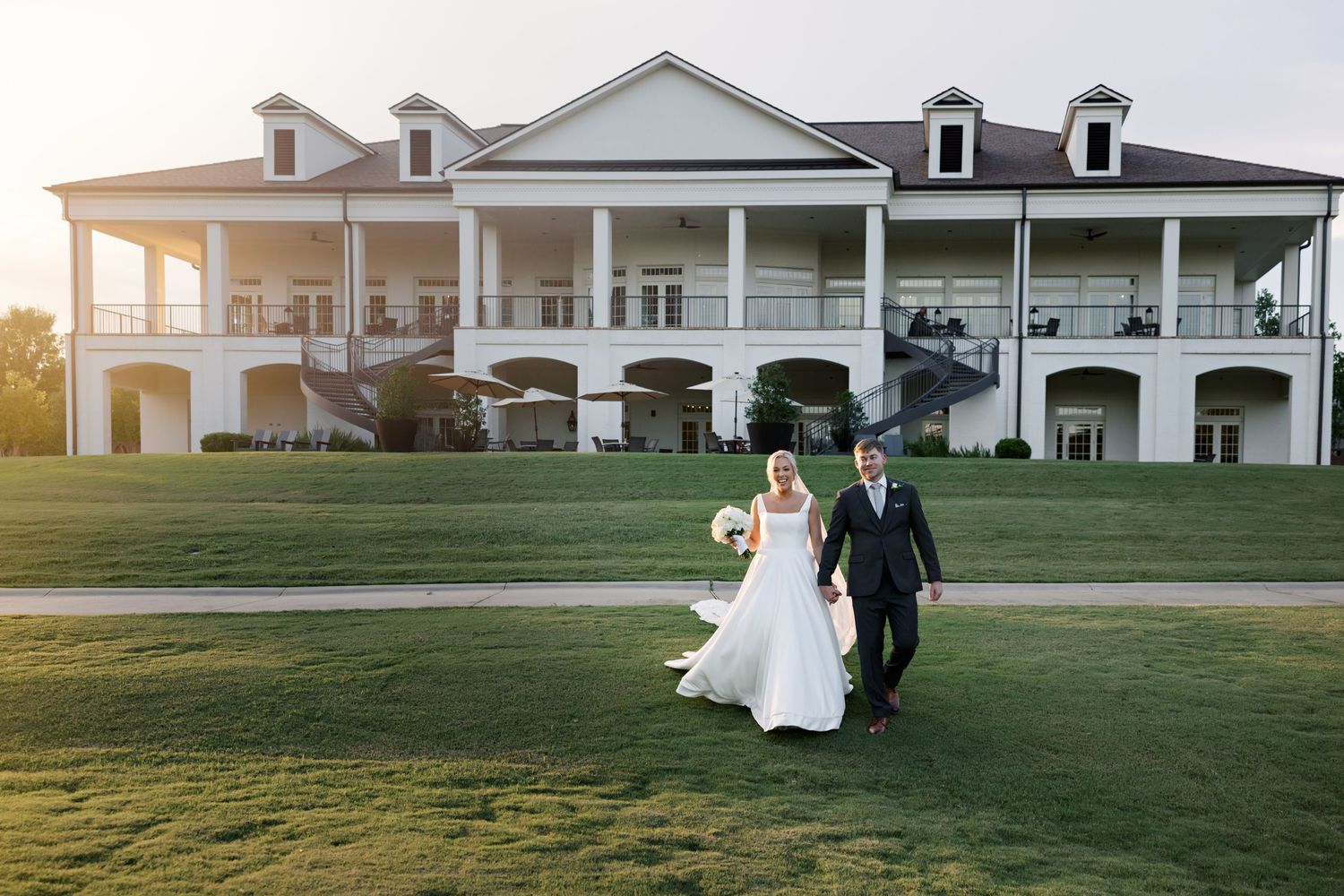 Couple in wedding attire walks together on a lush green lawn in front of a stately white mansion at sunset.