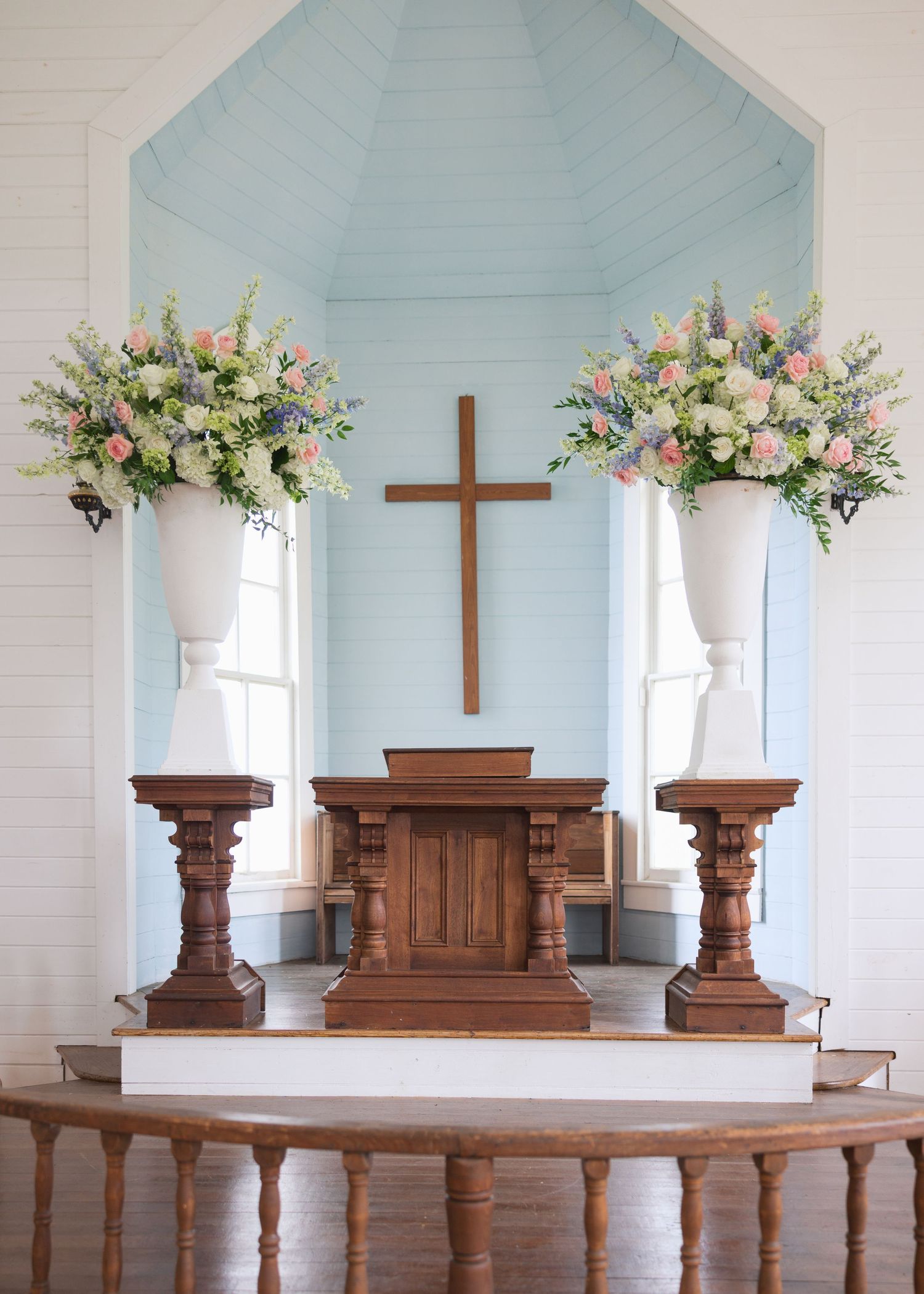 Church altar with wooden cross flanked by two large floral arrangements in soft pastel colors.
