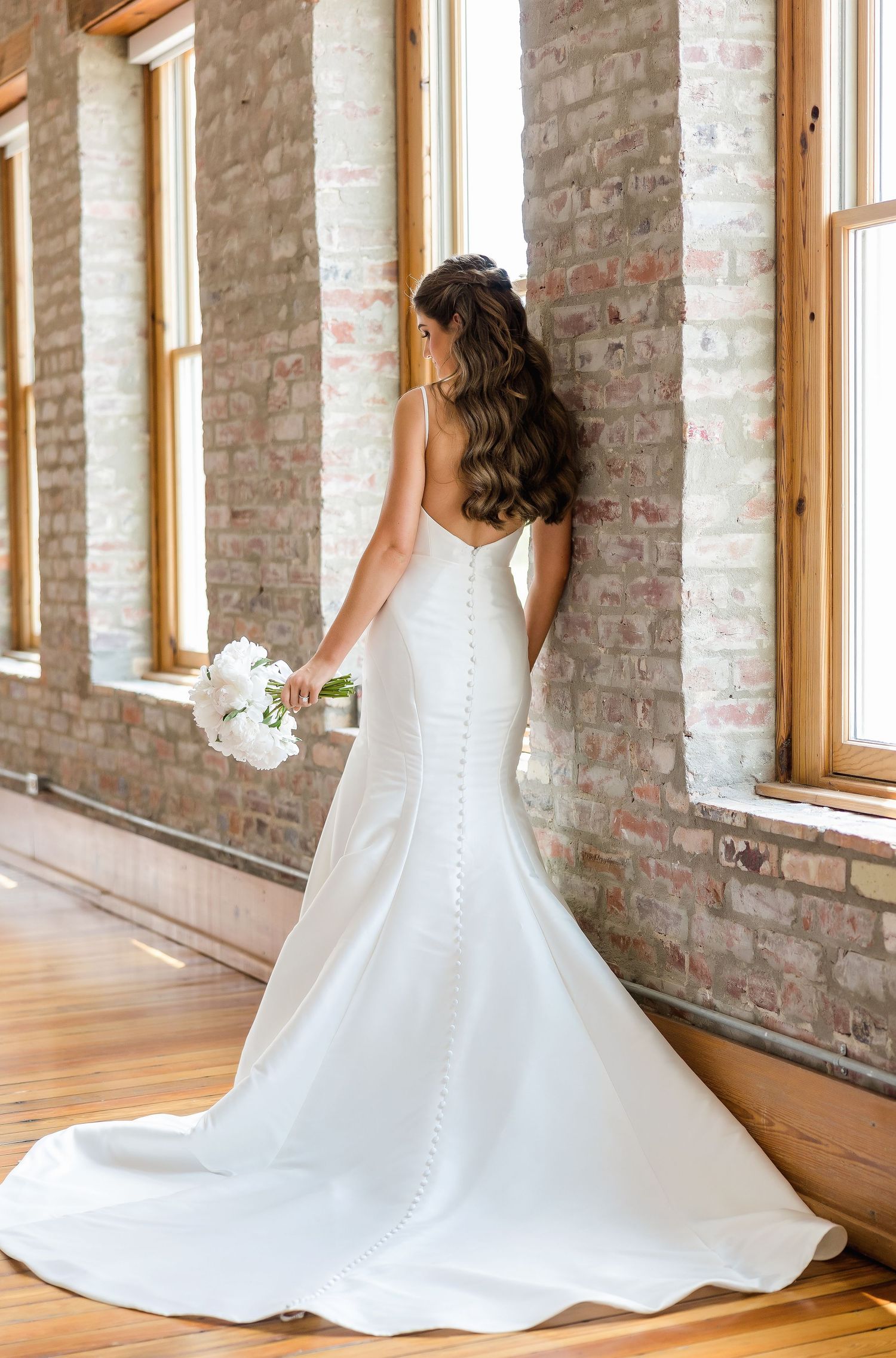 Bride in fitted white gown with long train poses by exposed brick windows.