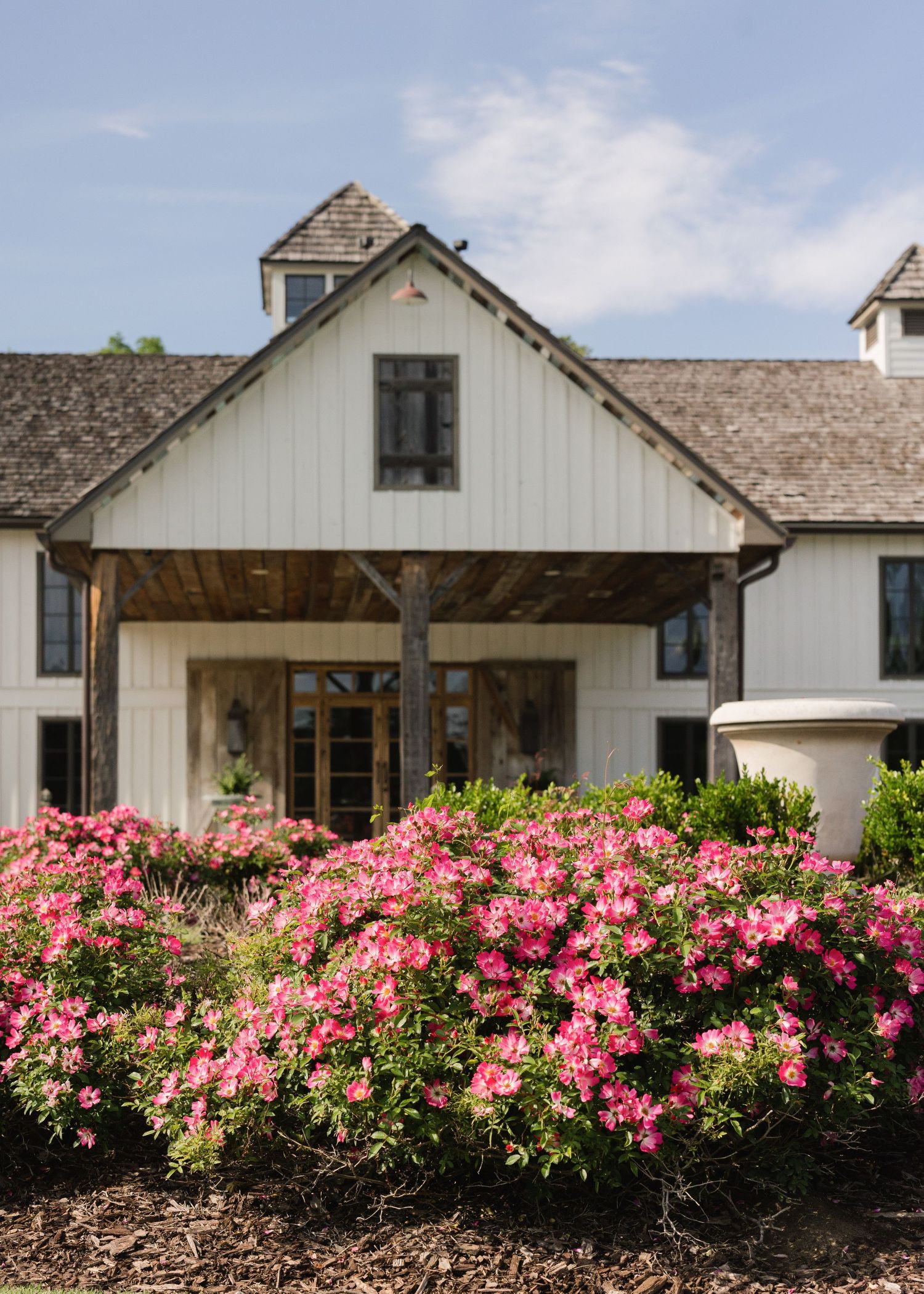 White farmhouse exterior with pink flowering bushes and rustic architecture.