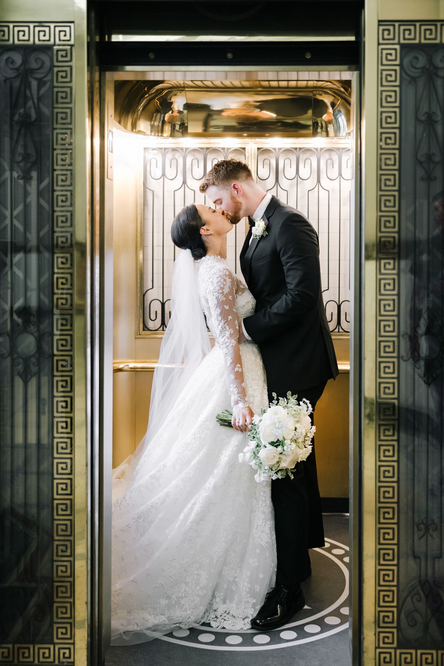 Elegant wedding couple sharing a kiss in an ornate gold elevator with art deco details.