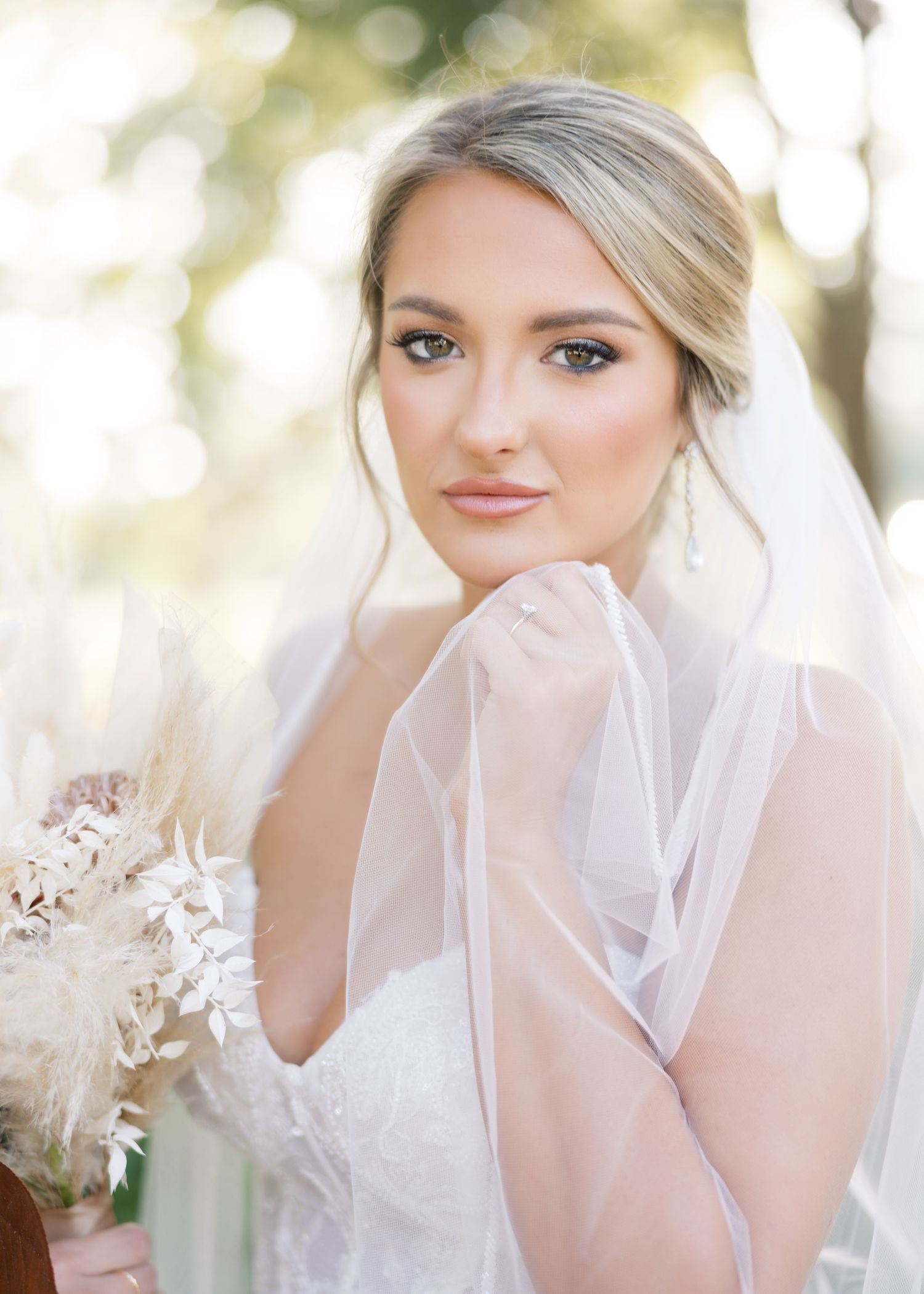Elegant bride in white gown and veil poses with natural light for portrait.