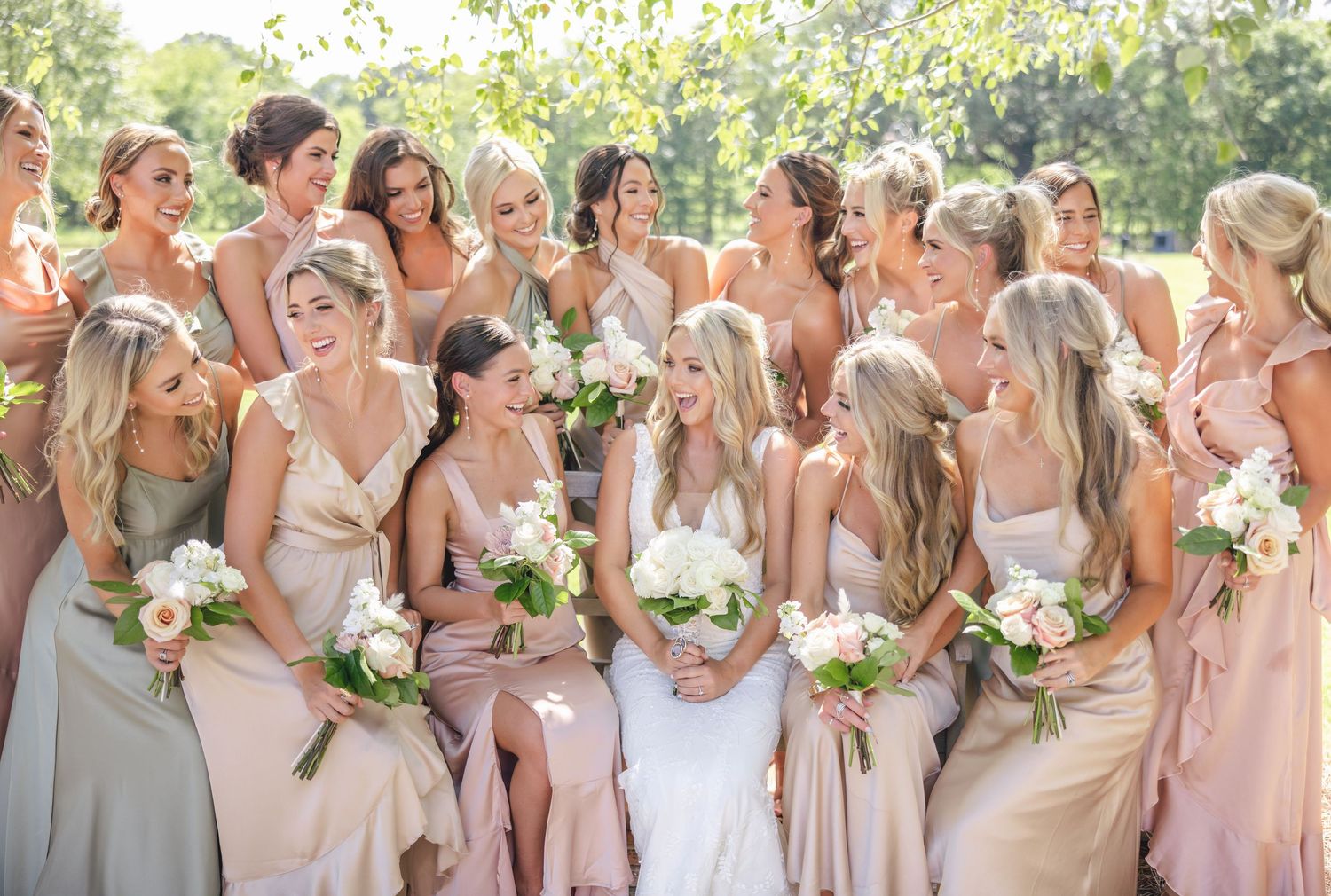 Large bridal party in blush pink dresses pose together outdoors with bouquets.