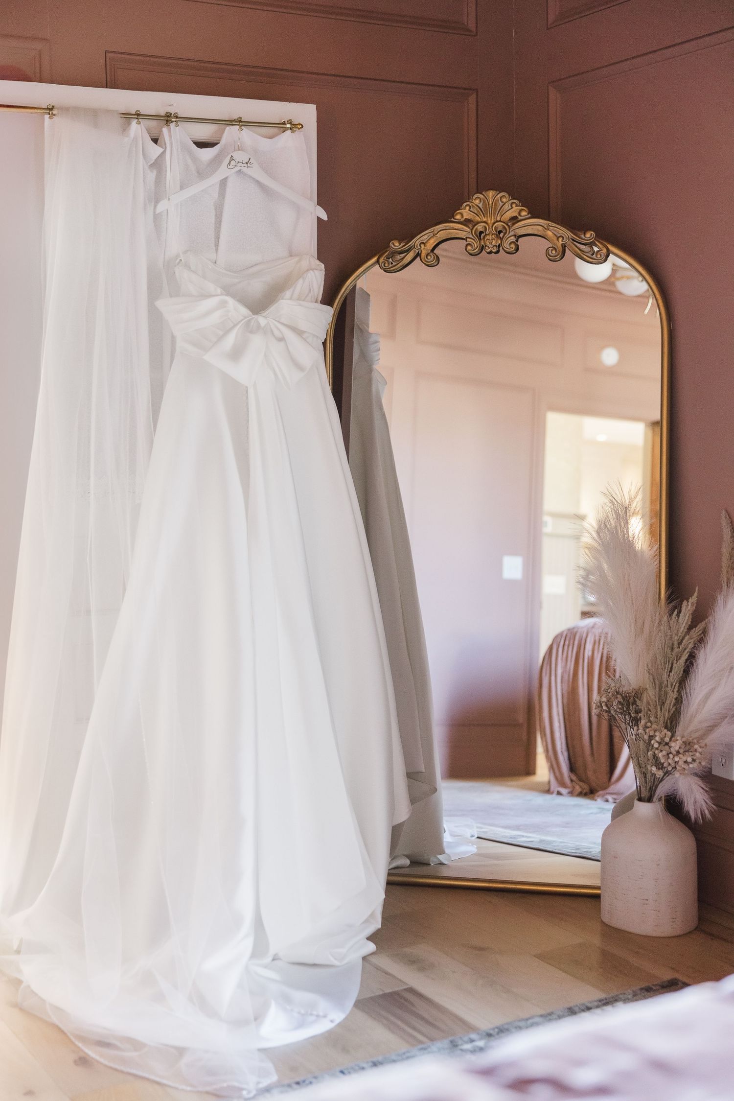 White wedding dress displayed on hangers with ornate mirror in soft pink room.