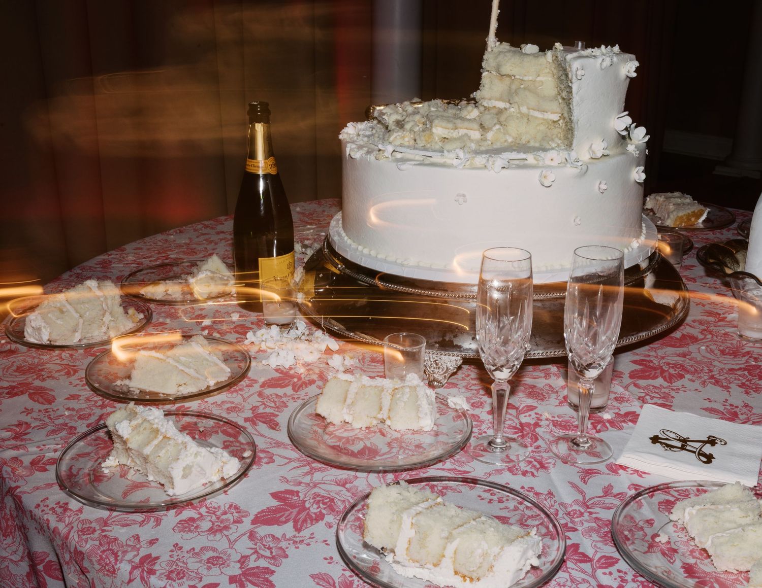 A white wedding cake with floral decorations sits on an elegant table with a pink patterned tablecloth.