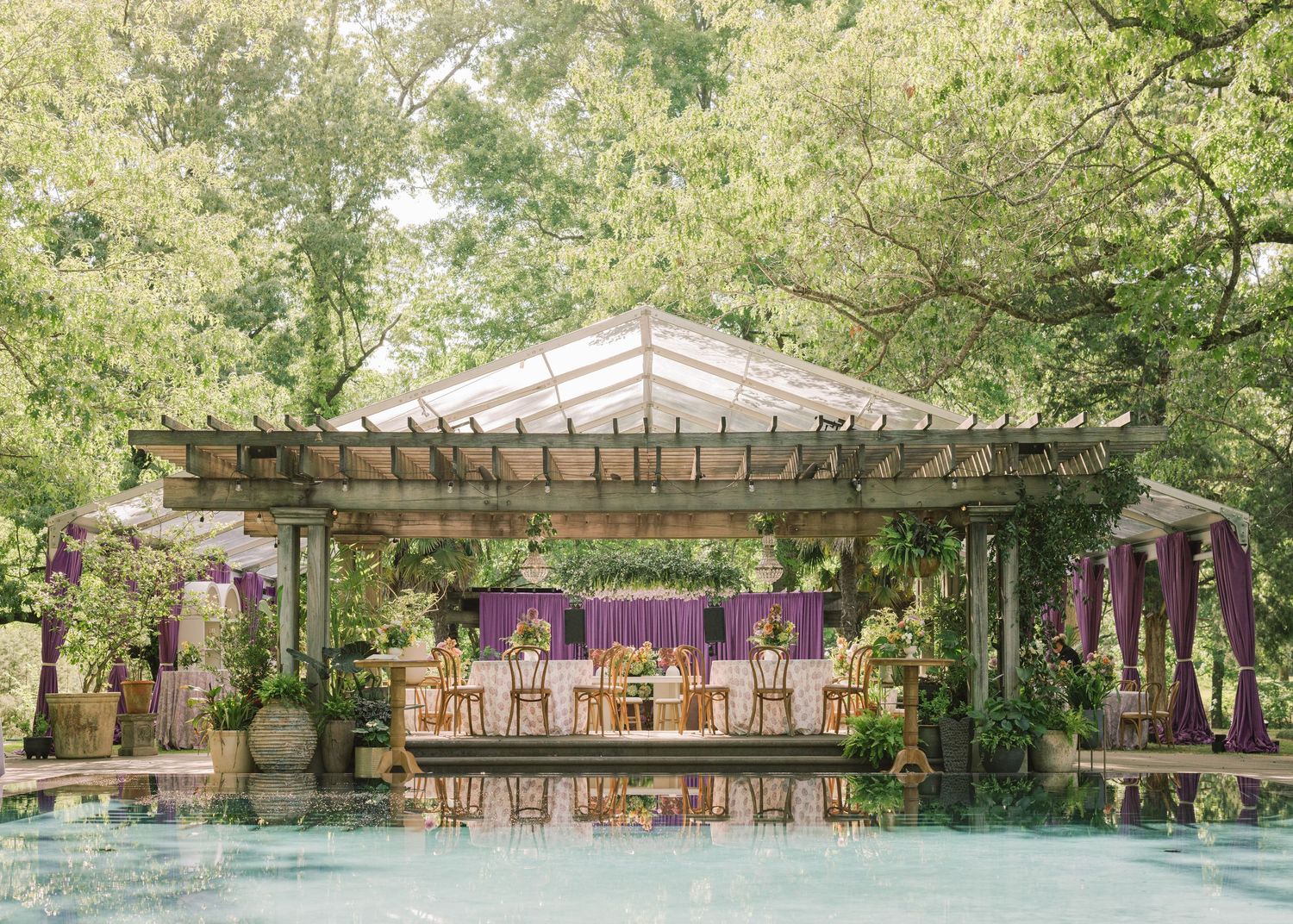 Elegant outdoor wedding reception pavilion adorned with purple drapes and floral arrangements beside a serene pool.