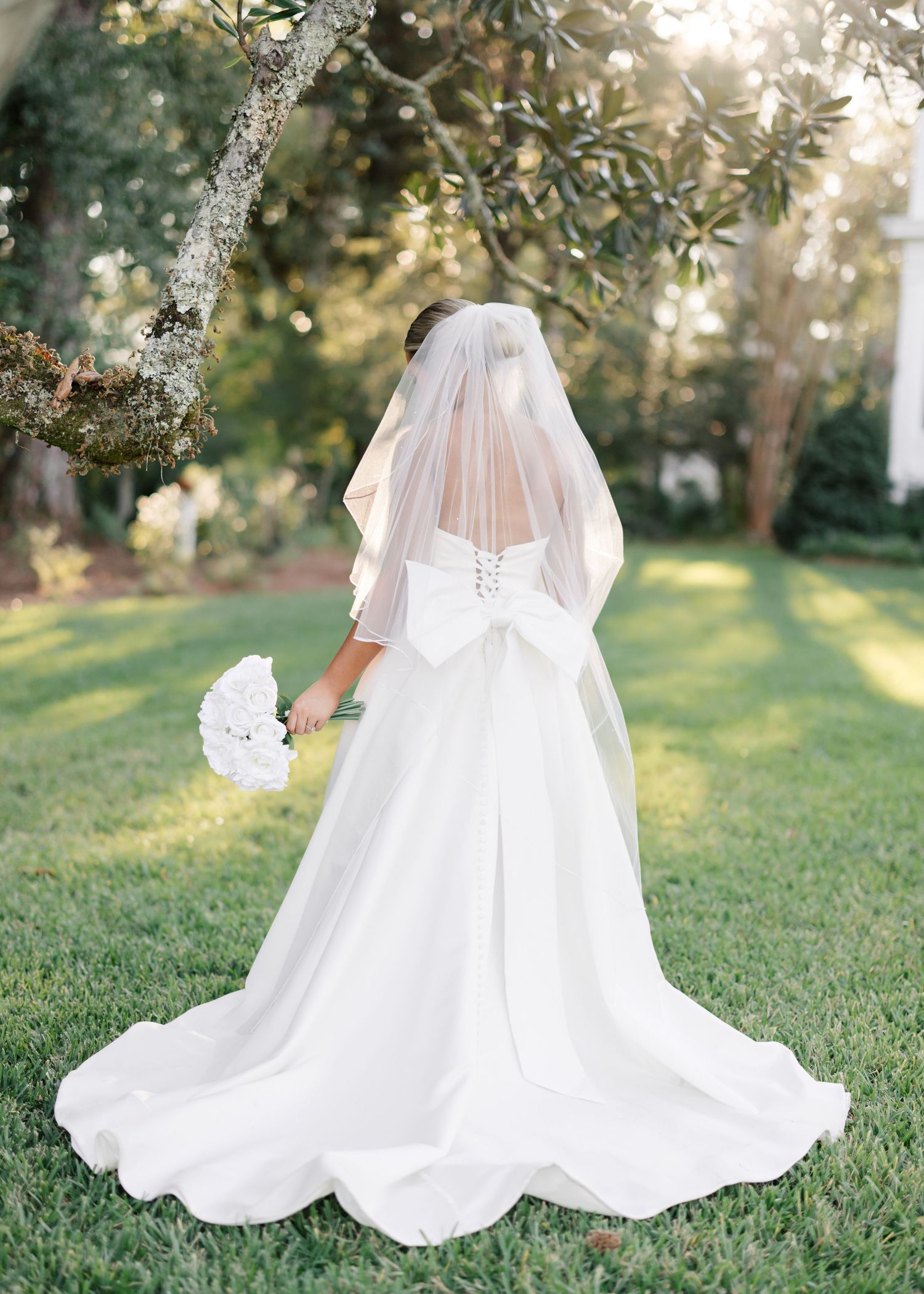 Flowing white wedding dress with cape details twirls gracefully on green grass under sunlit trees.