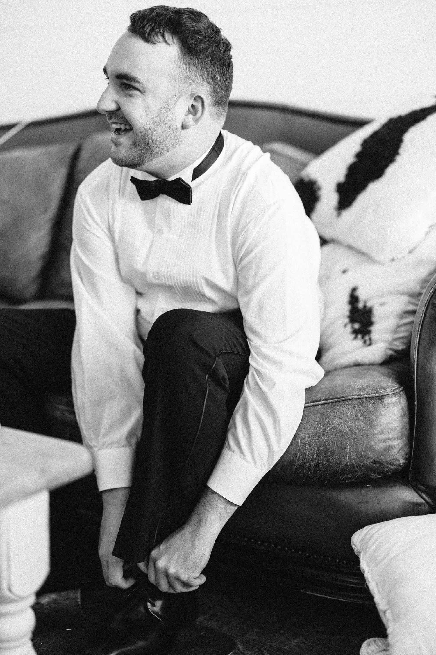Person in white dress shirt and black vest sitting on leather couch smiling in black and white photo.