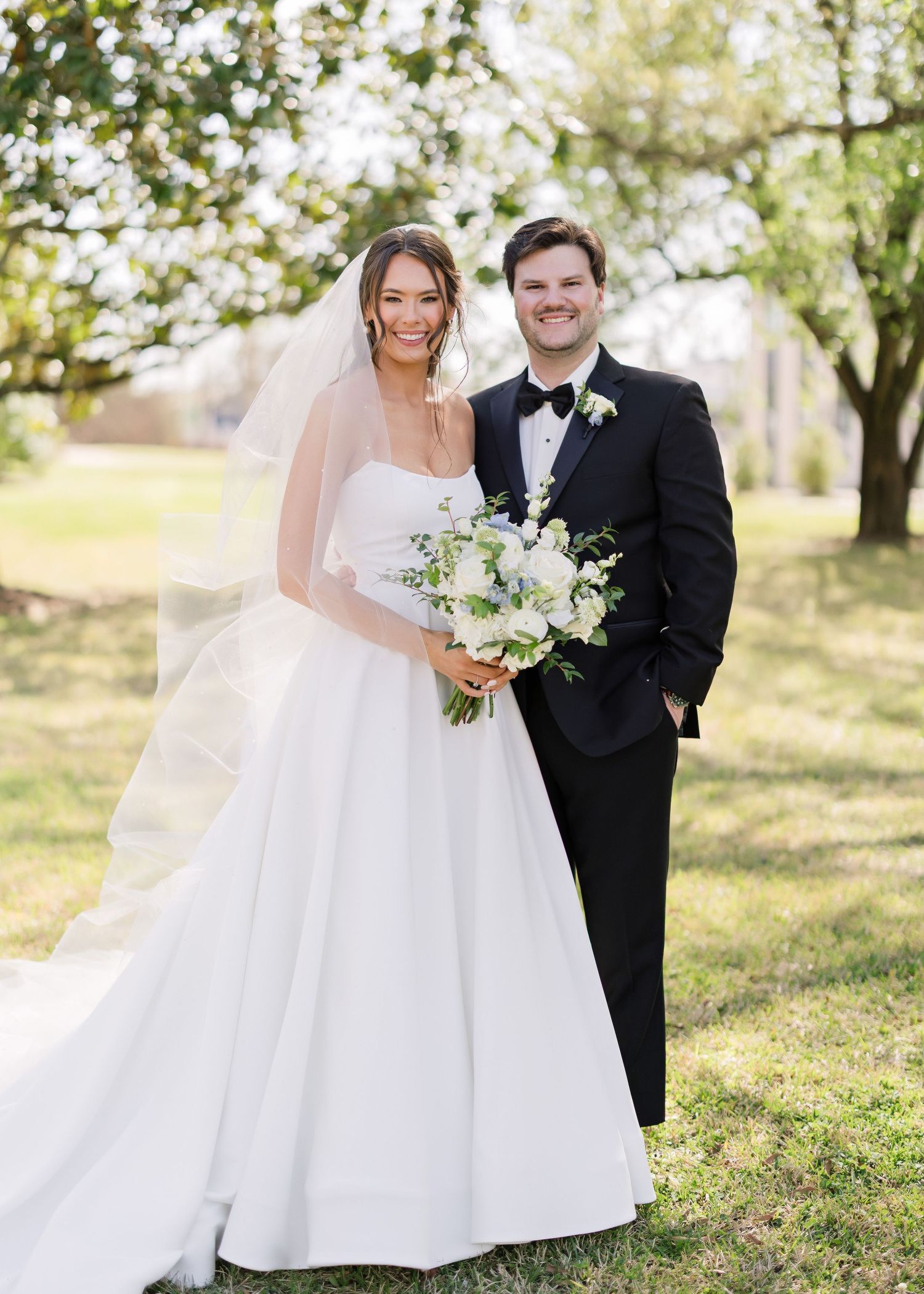 Wedding couple poses lovingly together on sunny grass with classic white gown and black tuxedo.