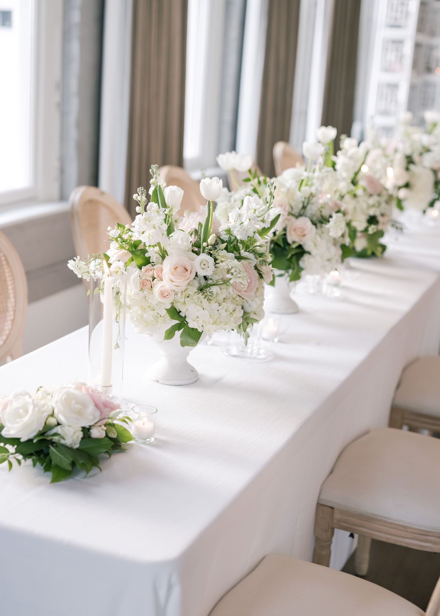 Elegant white and blush floral centerpieces line a wedding reception table with white linens.