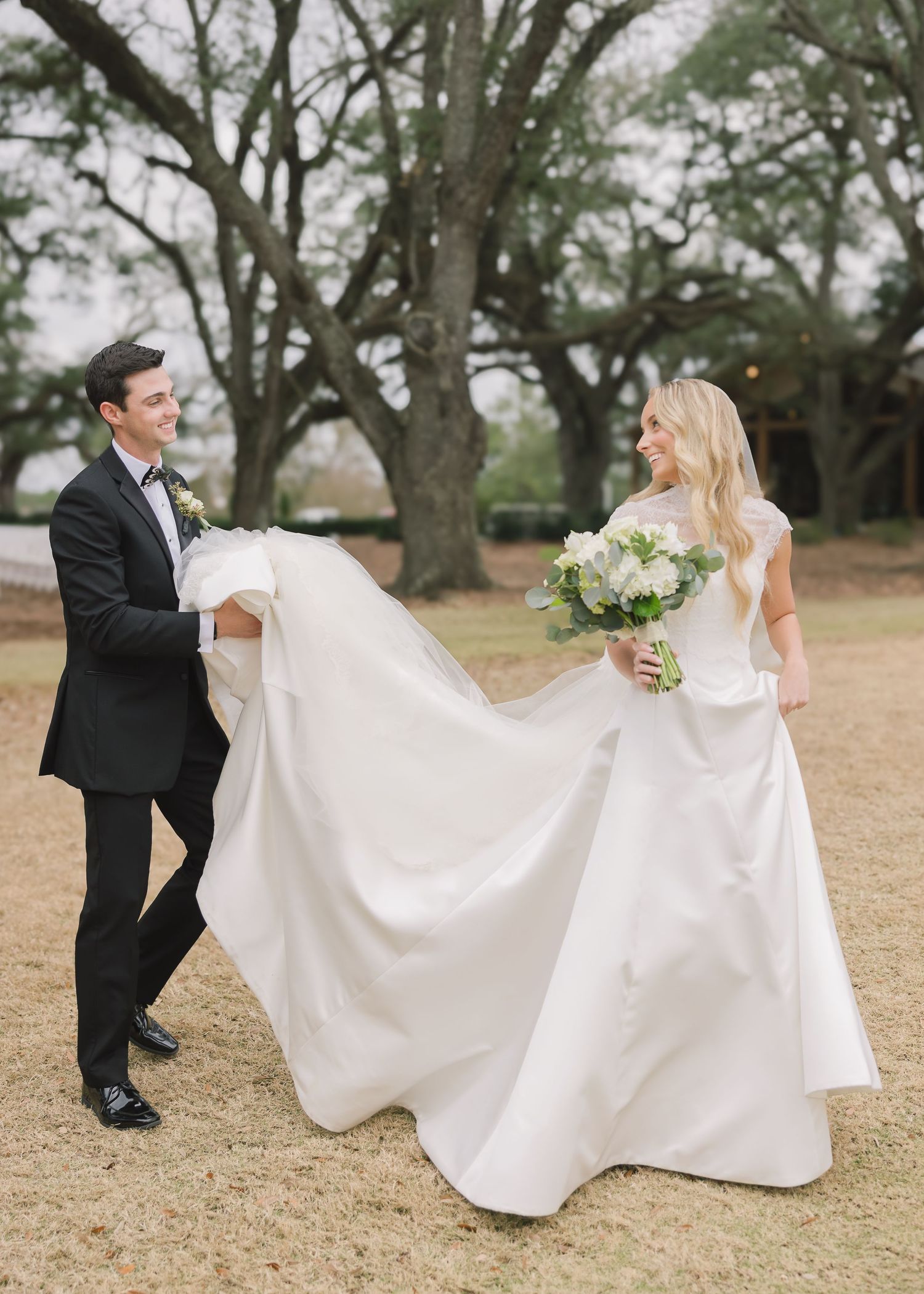 A couple in wedding attire playfully walks through a grassy field as the groom helps carry the bride's flowing dress train.