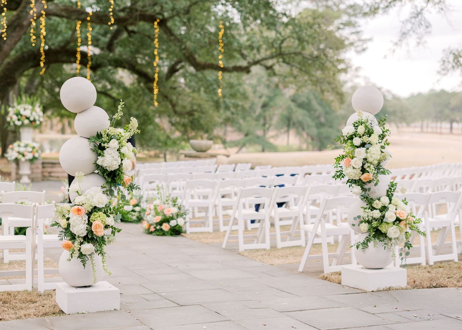 Elegant outdoor wedding ceremony setup with white chairs and floral arrangements along a tree-lined aisle.