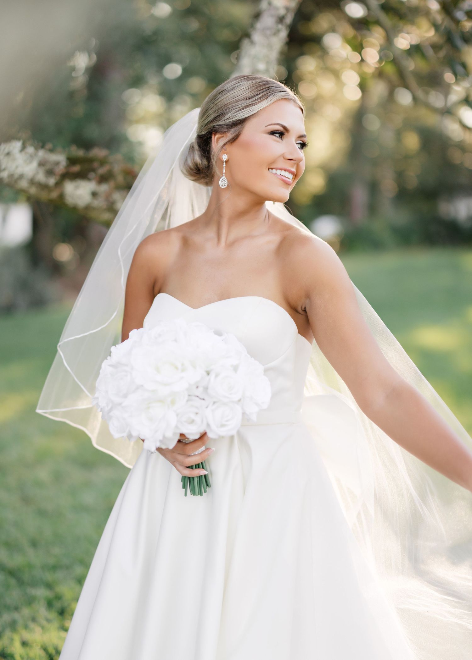 A bride in a strapless white wedding gown poses outdoors in natural sunlight with a flowing veil.