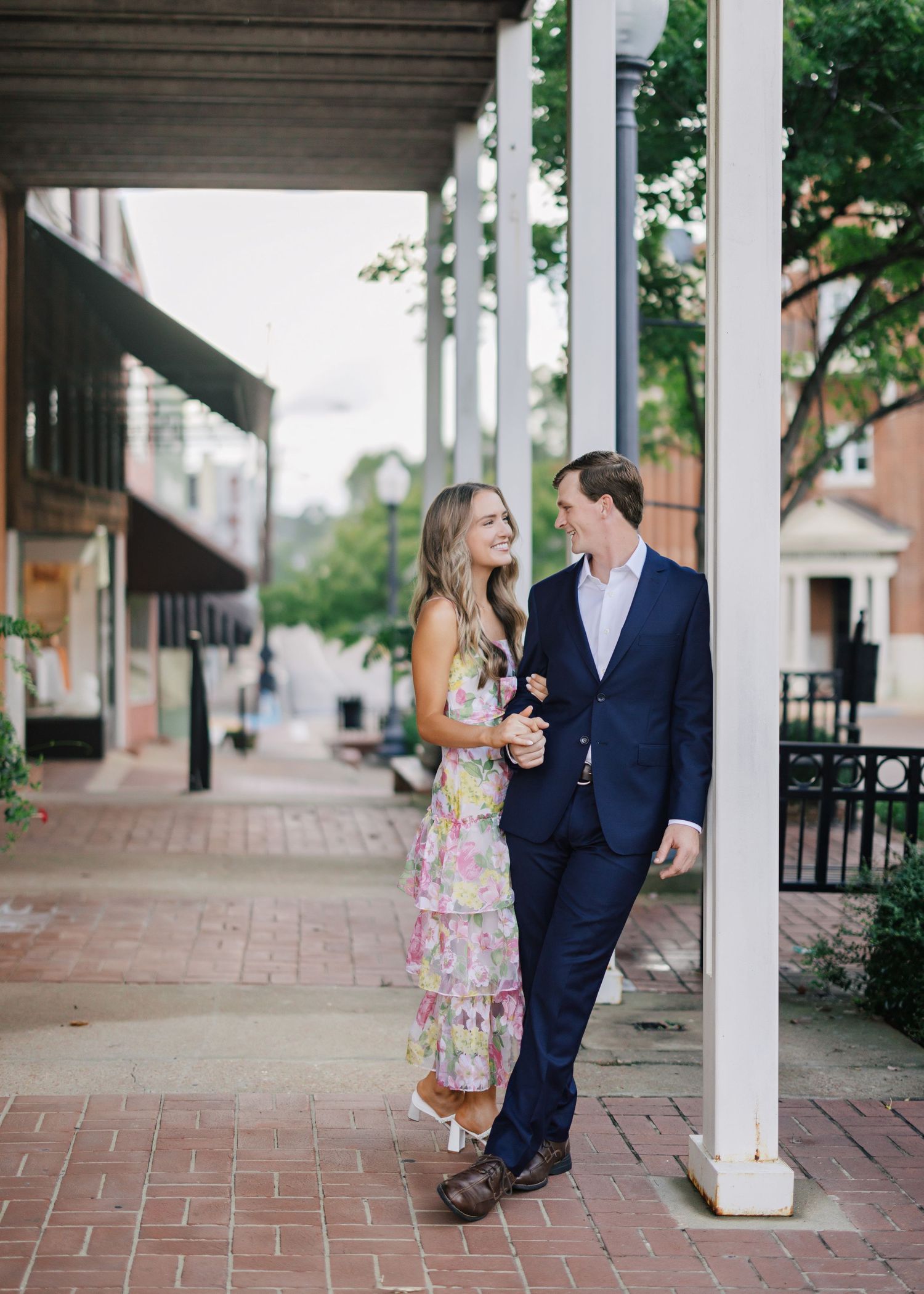 Couple walking together on a brick sidewalk with white columns, with her in a floral dress and him in a navy suit.