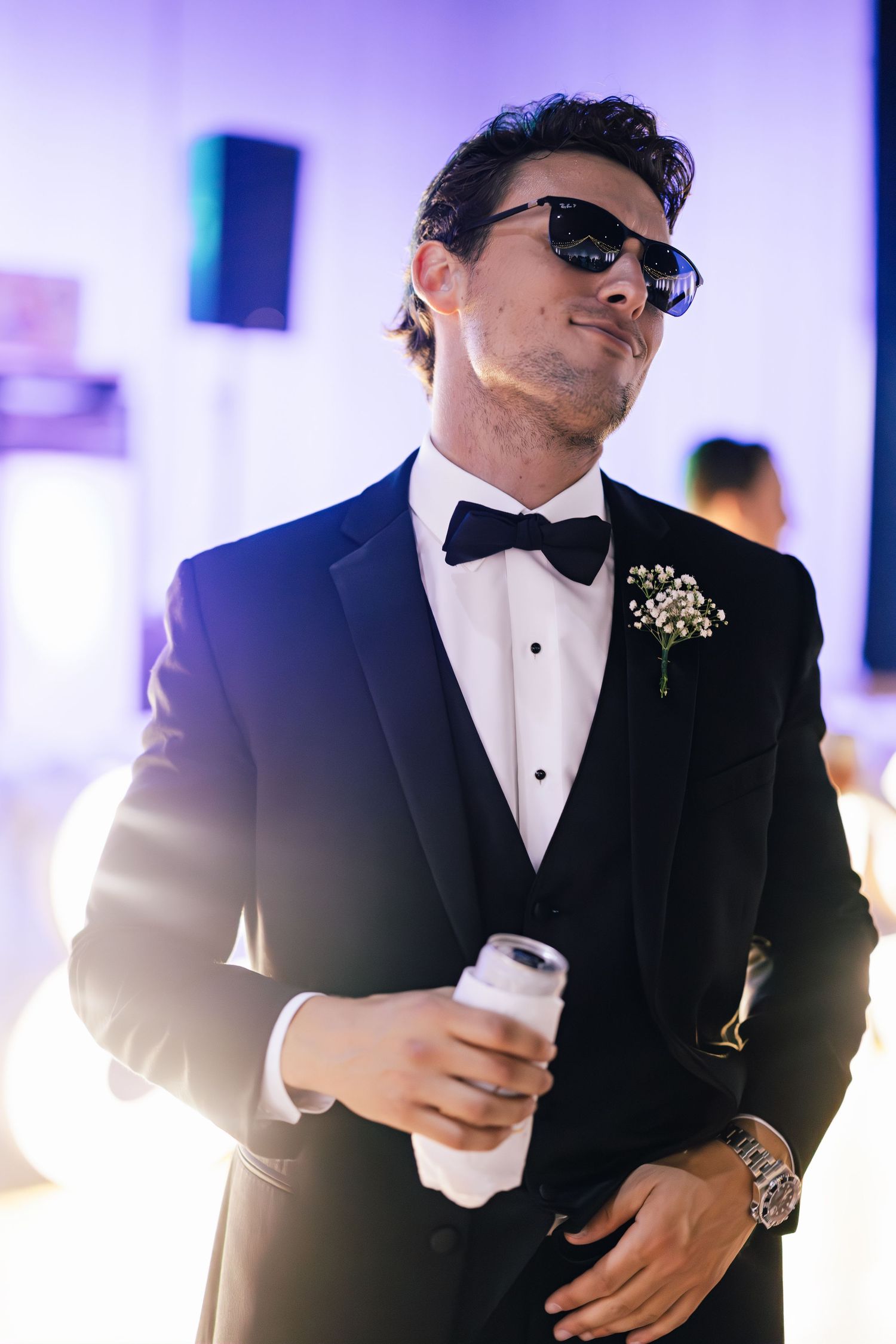 Groomsmen in black tuxedos and sunglasses posing with drinks at reception.