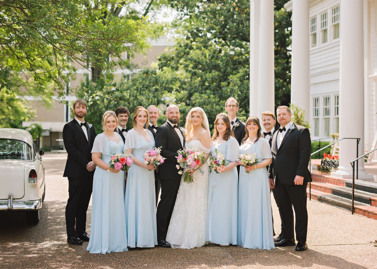 Wedding party in light blue bridesmaid dresses and formal attire gathered on outdoor porch.