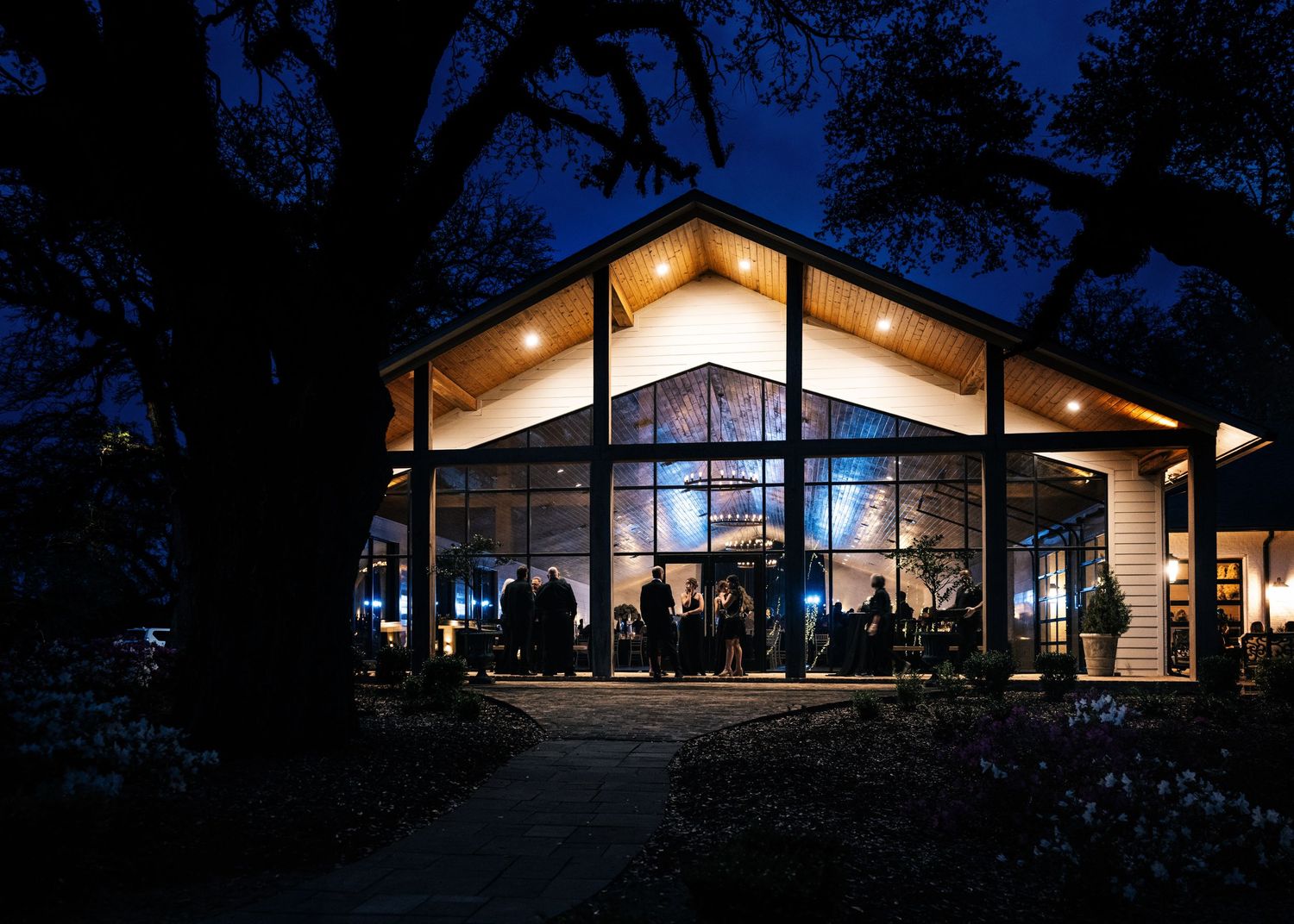 Modern glass wedding venue glowing at twilight with silhouettes of guests inside.