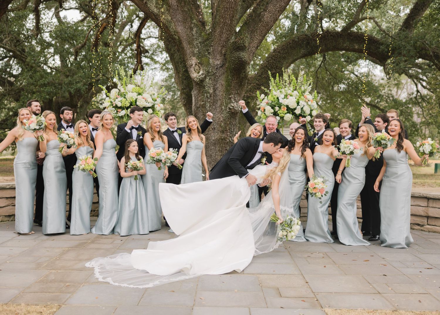 A wedding party in light blue bridesmaid dresses poses together under large oak trees on a stone patio.