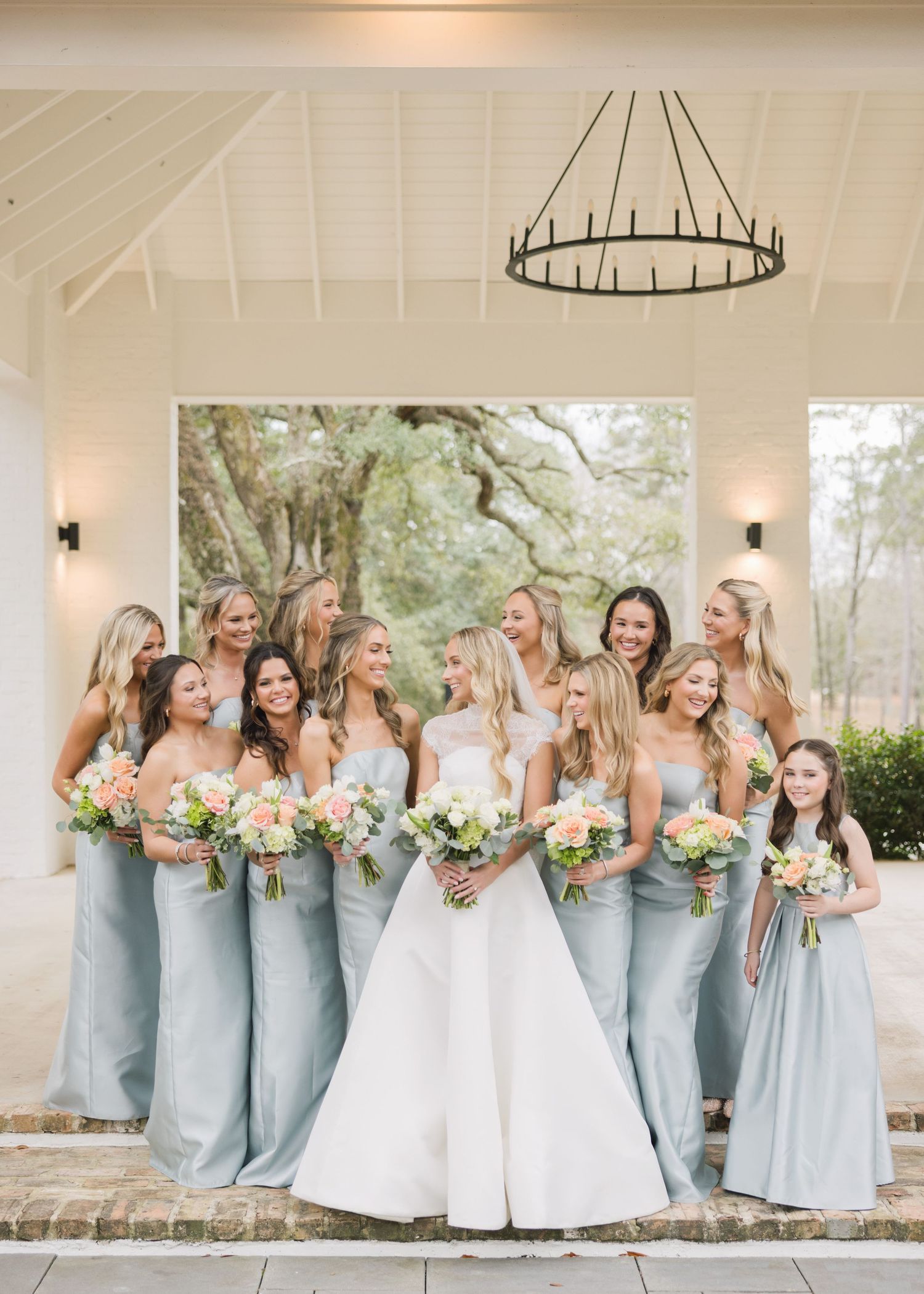 A group of bridesmaids in light blue dresses pose together with the bride in white at an outdoor wedding venue.