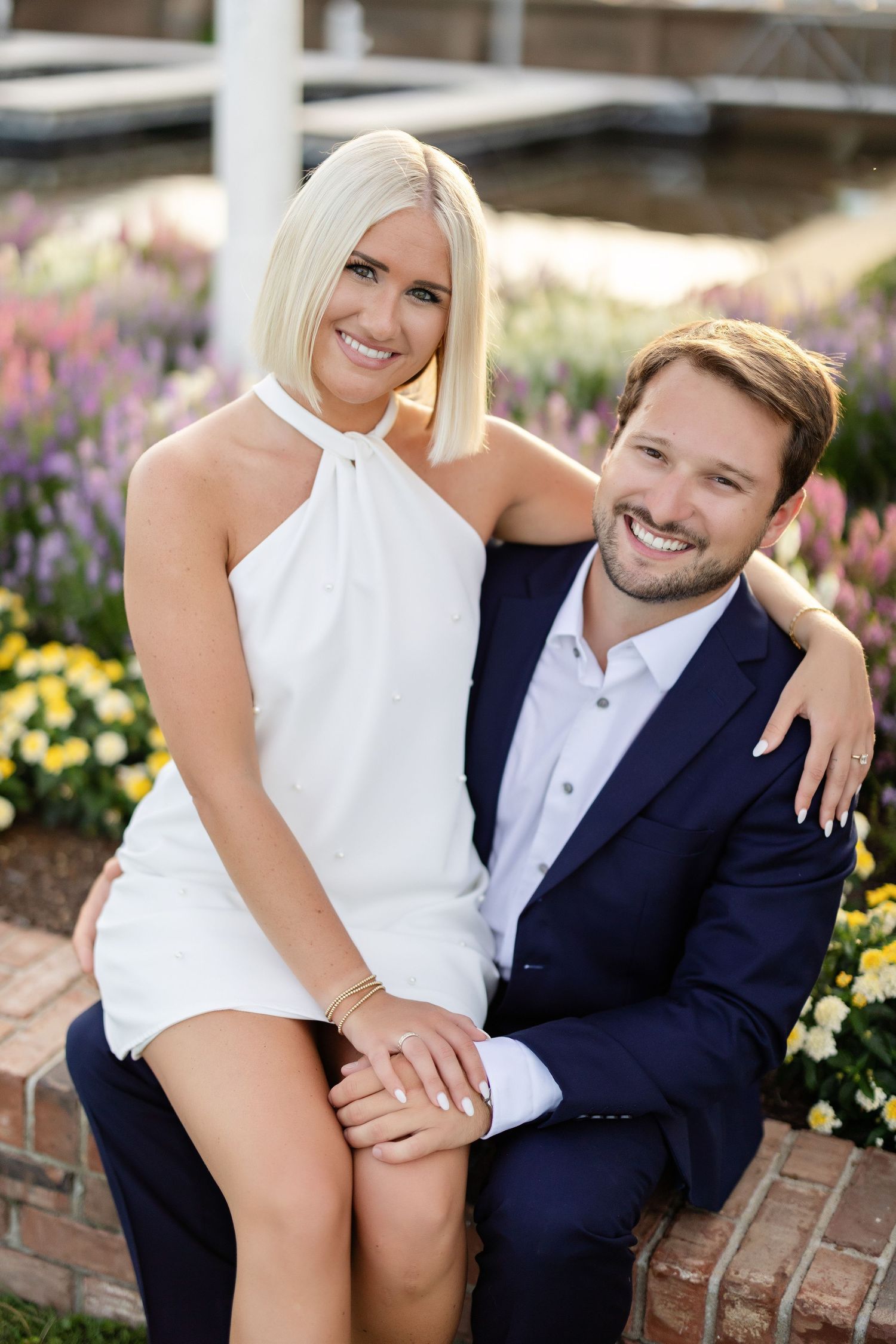 Couple posing together in front of colorful garden flowers with woman in white dress and man in navy suit.