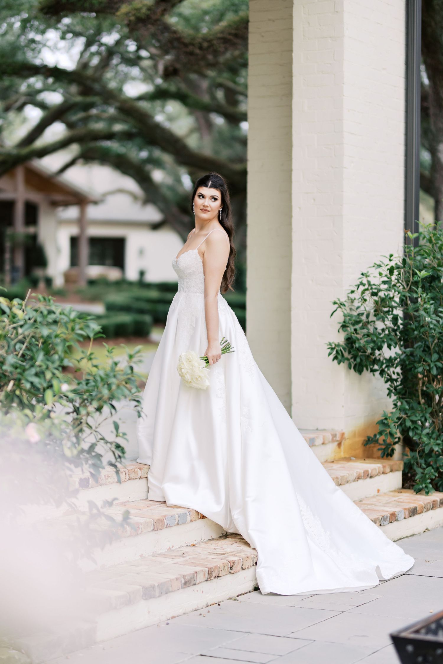 Elegant bride in white strapless gown poses on stone steps near garden pillars.