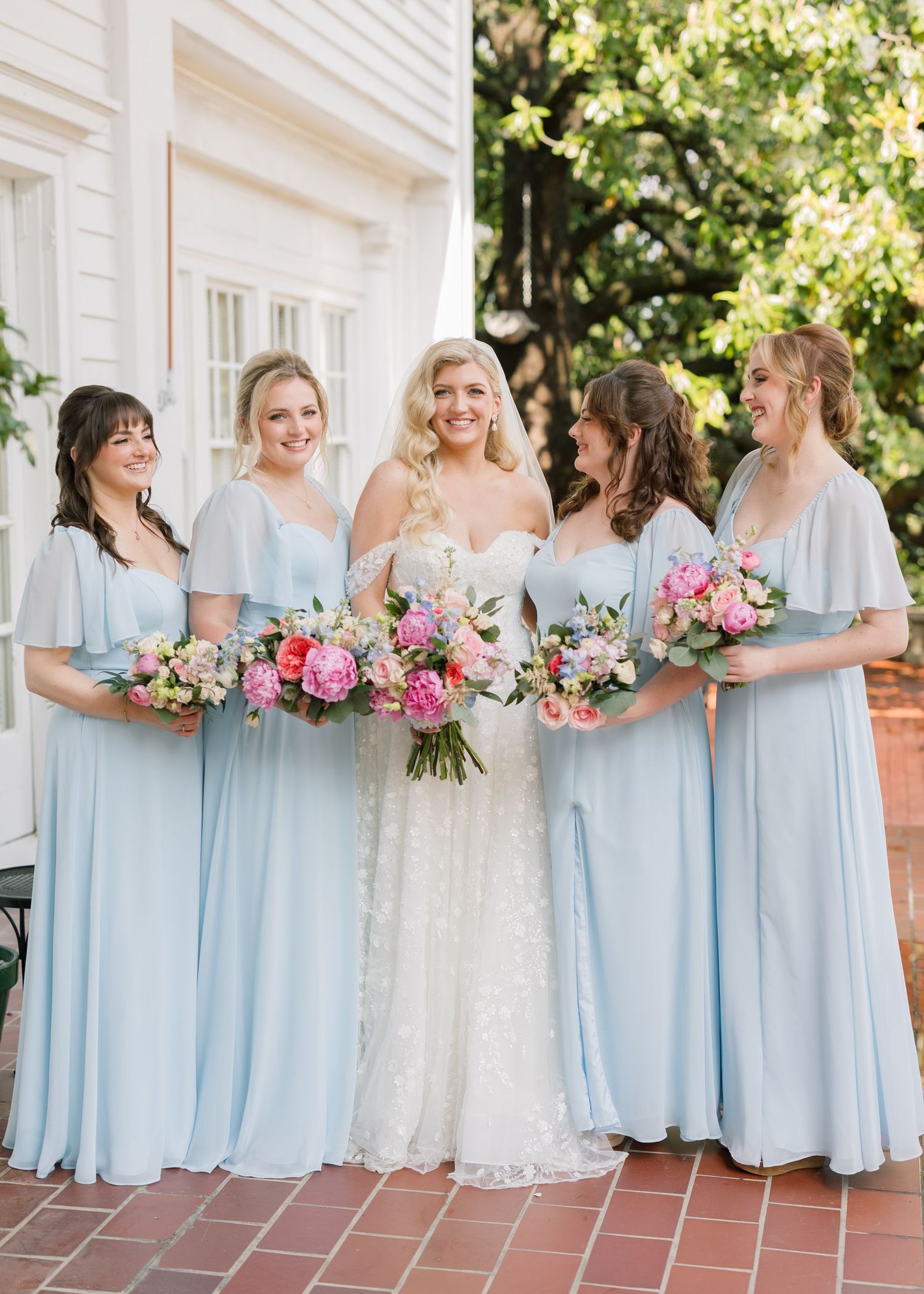 Bridesmaids in light blue dresses holding pink and white bouquets on a brick porch.