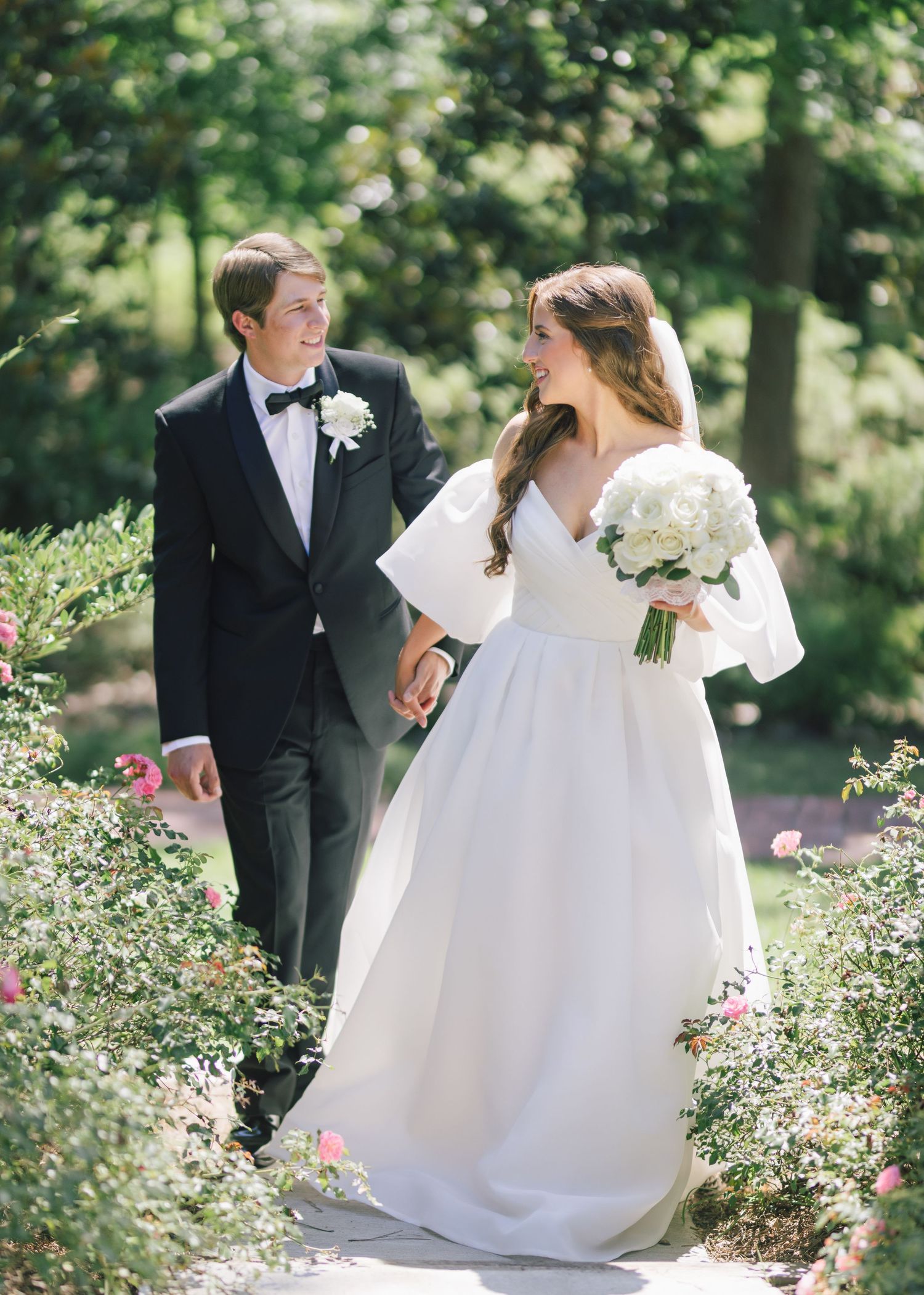 A couple walks through a garden pathway surrounded by blooming flowers on their wedding day.