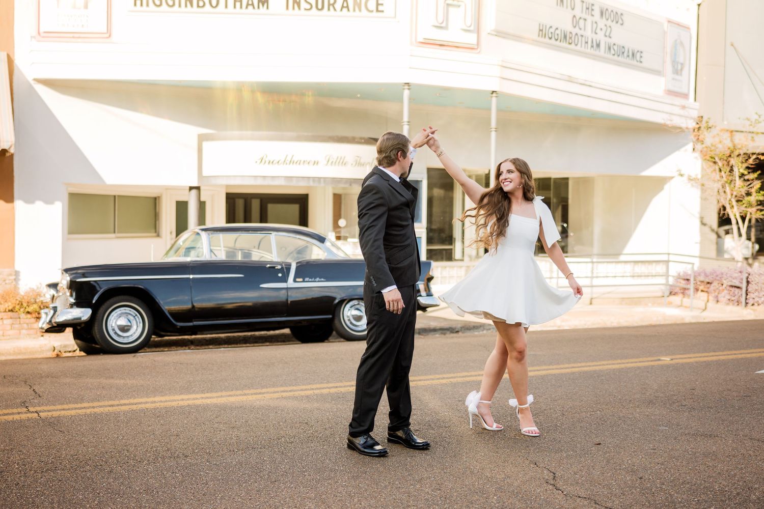 Wide angle engagement photo featuring couple and vintage car outside a storefront.