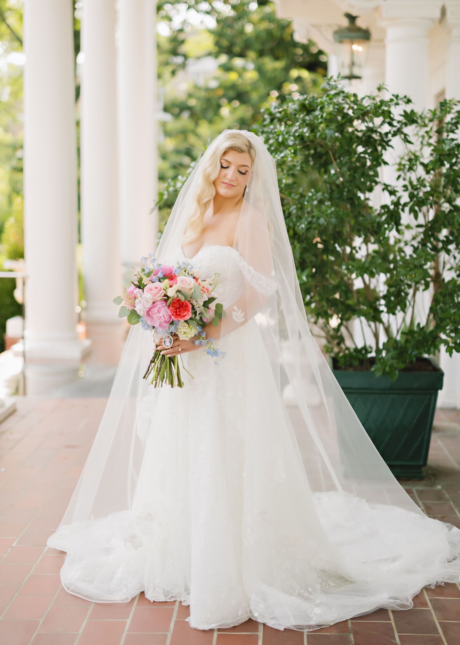 Bride in a flowing white wedding dress and veil poses elegantly on a columned porch with pink and coral bouquet.