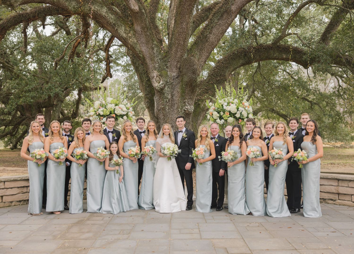 Wedding party in light blue dresses and suits poses together under oak trees in an outdoor setting.