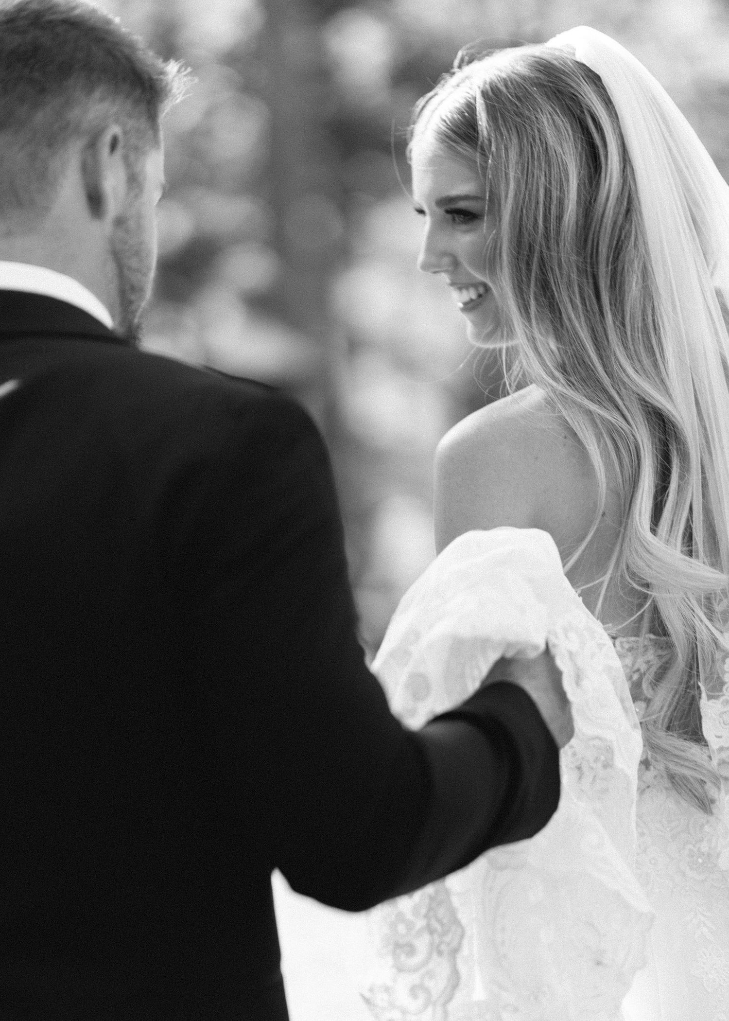 A bride in a white lace wedding dress laughs joyfully during an outdoor wedding ceremony.
