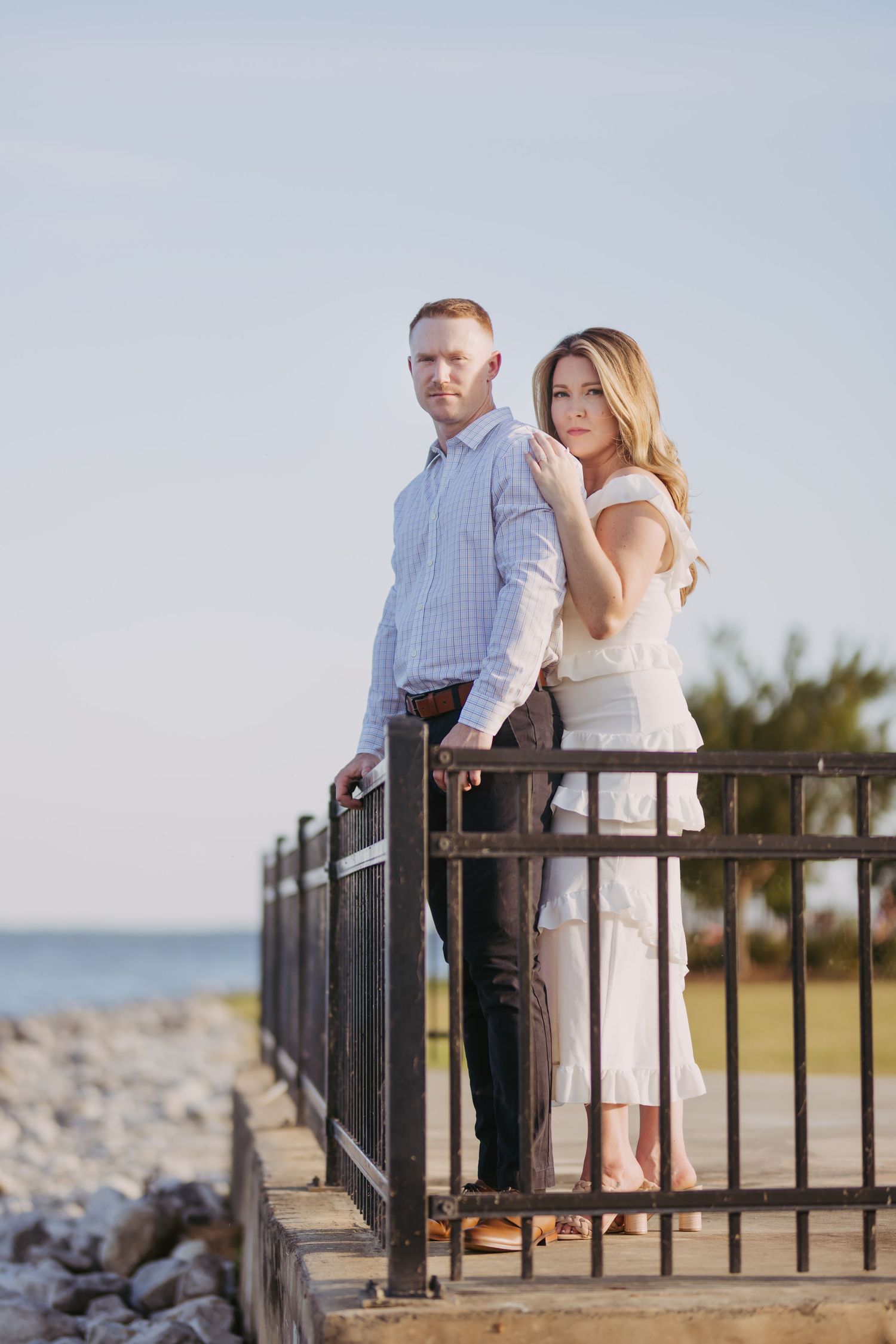Couple sharing romantic moment by metal railing with water and rocks in background.