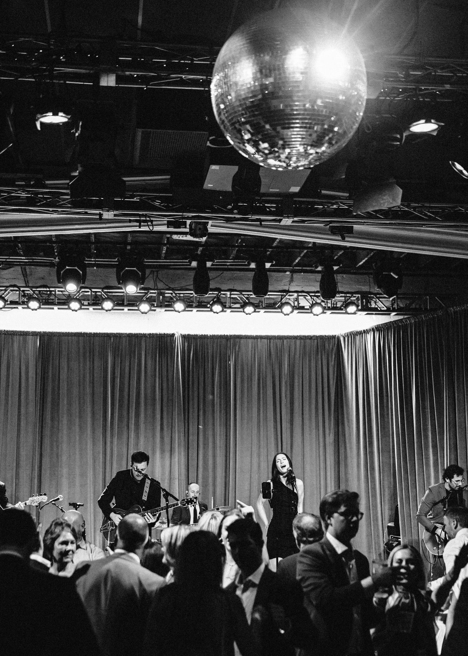 Large disco ball hangs over crowded dance floor at wedding reception with live band performing.