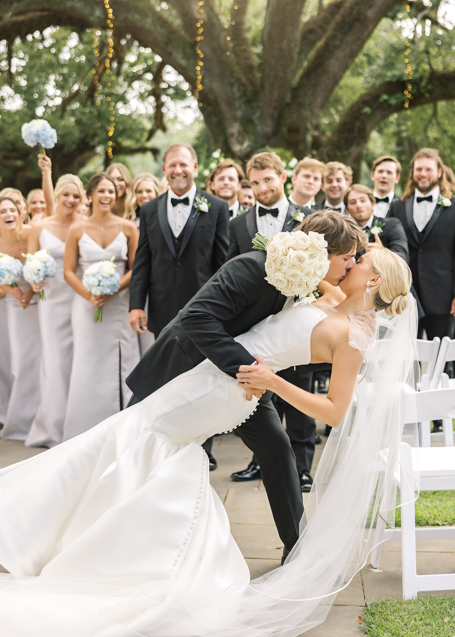 Wedding party celebrates with dips and poses under oak trees at outdoor ceremony.