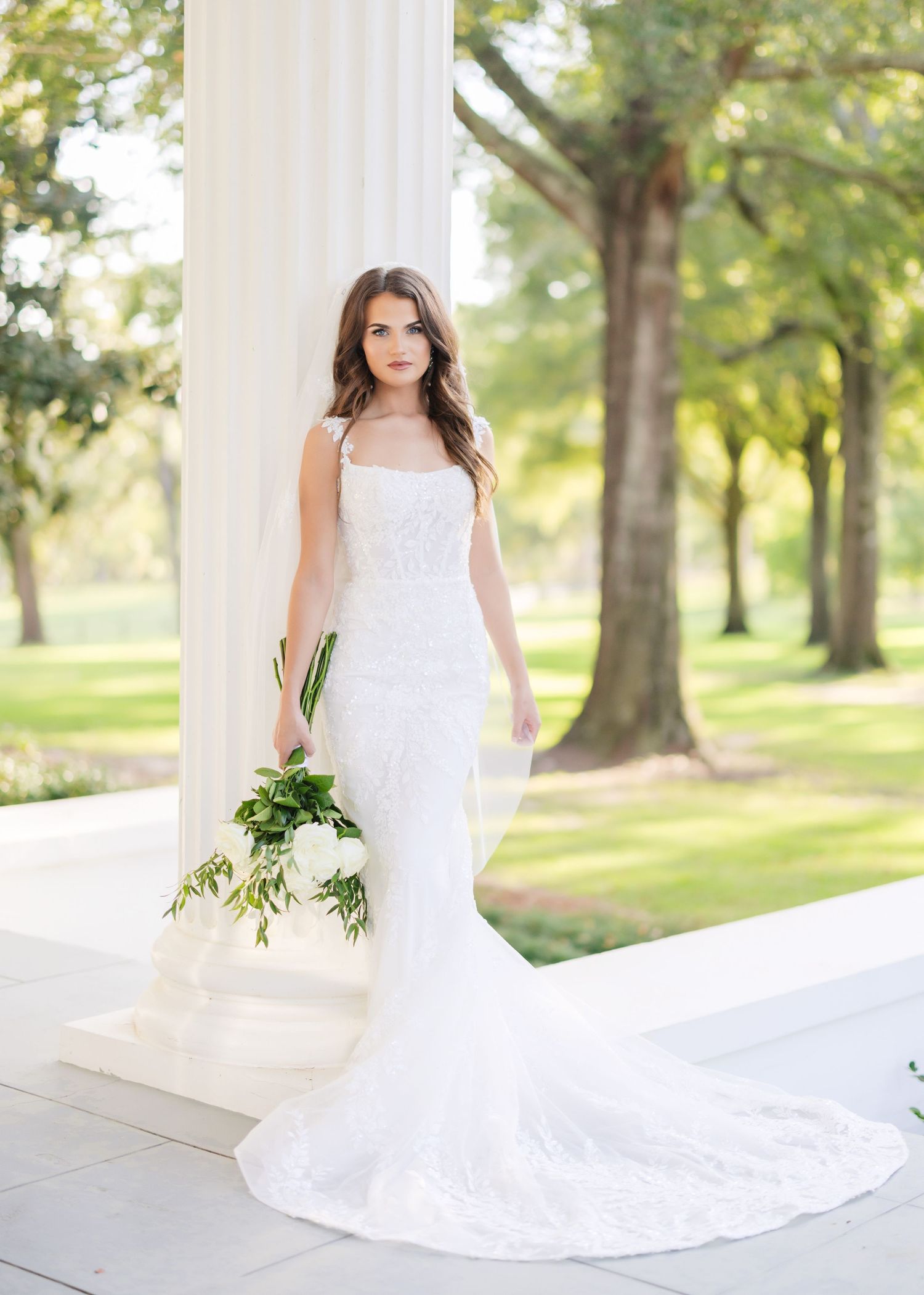 Elegant white bridal gown with train displayed against green garden pillars in outdoor setting.