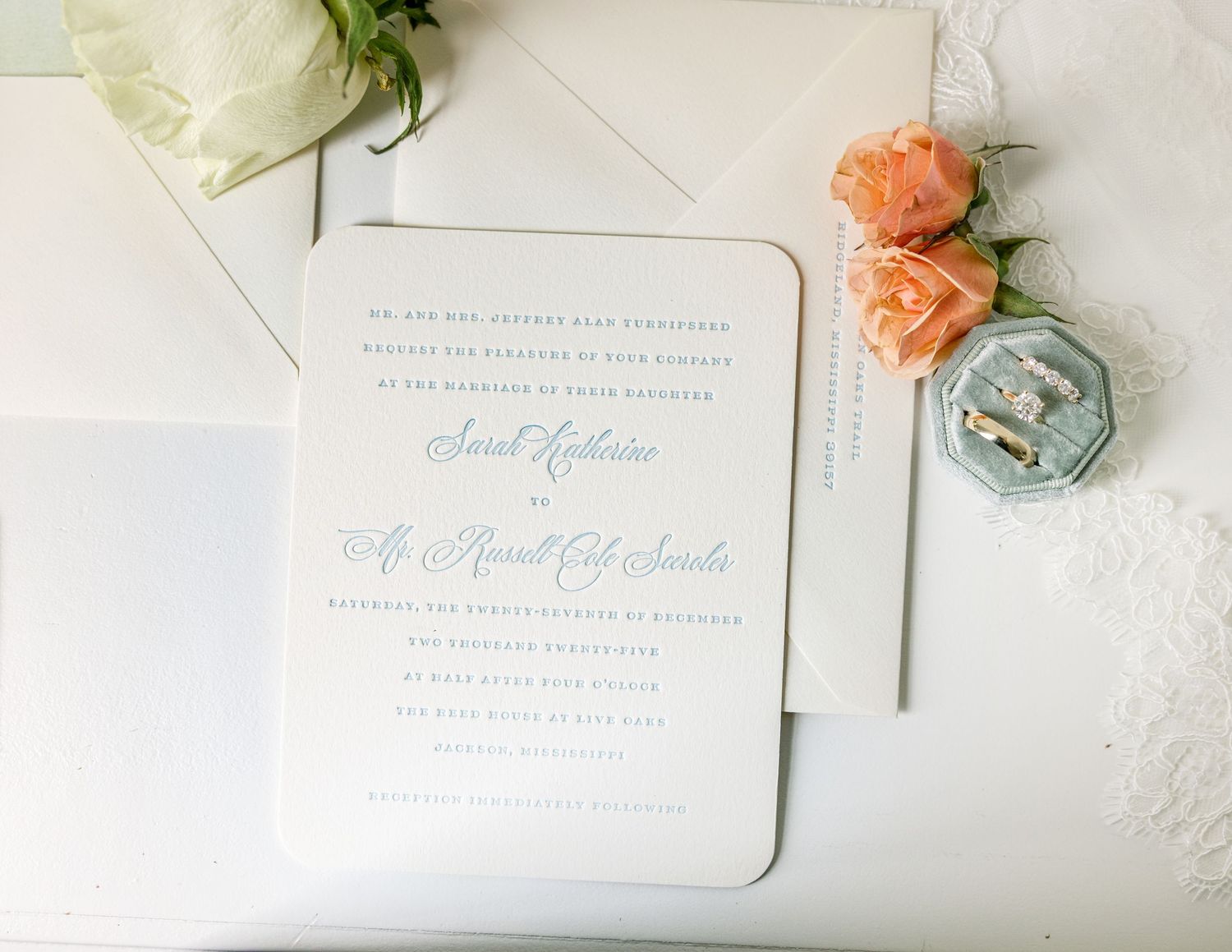 Elegant wedding invitation with pale blue text displayed alongside peach roses and a vintage-style ring box.