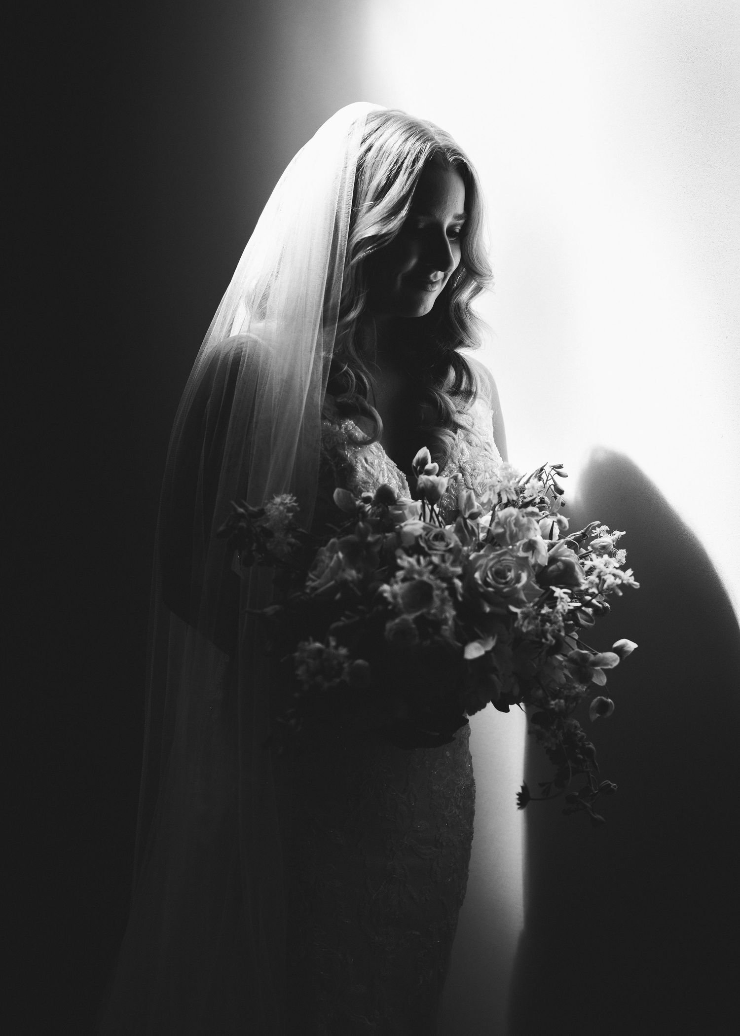 Dramatic black and white portrait of a bride holding a bouquet in soft lighting.