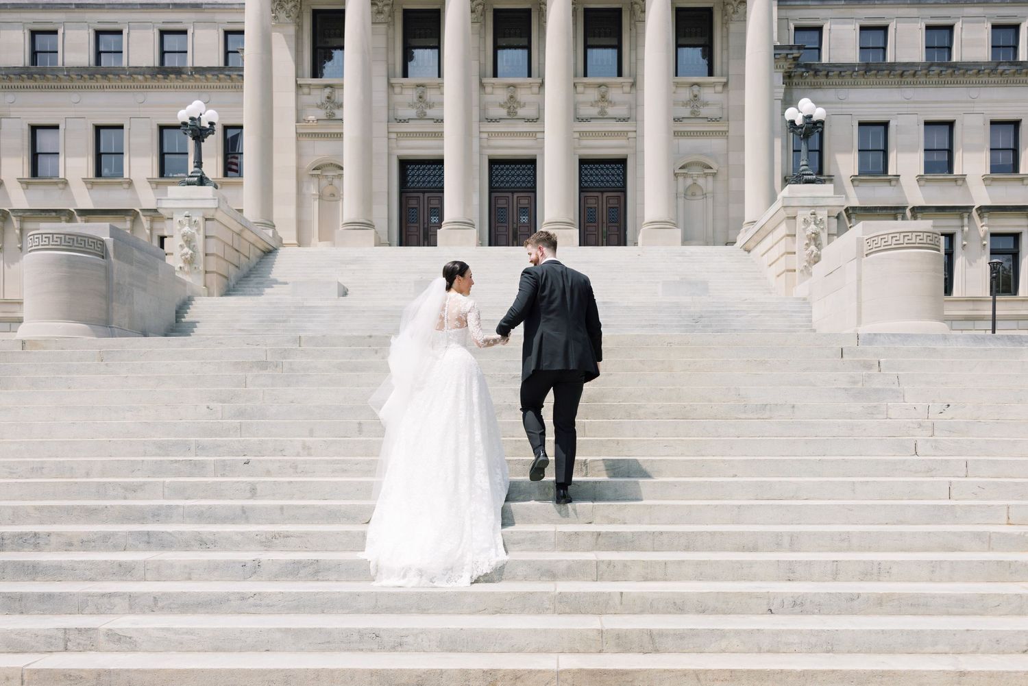 A couple ascends stone steps of a grand classical building on their wedding day.