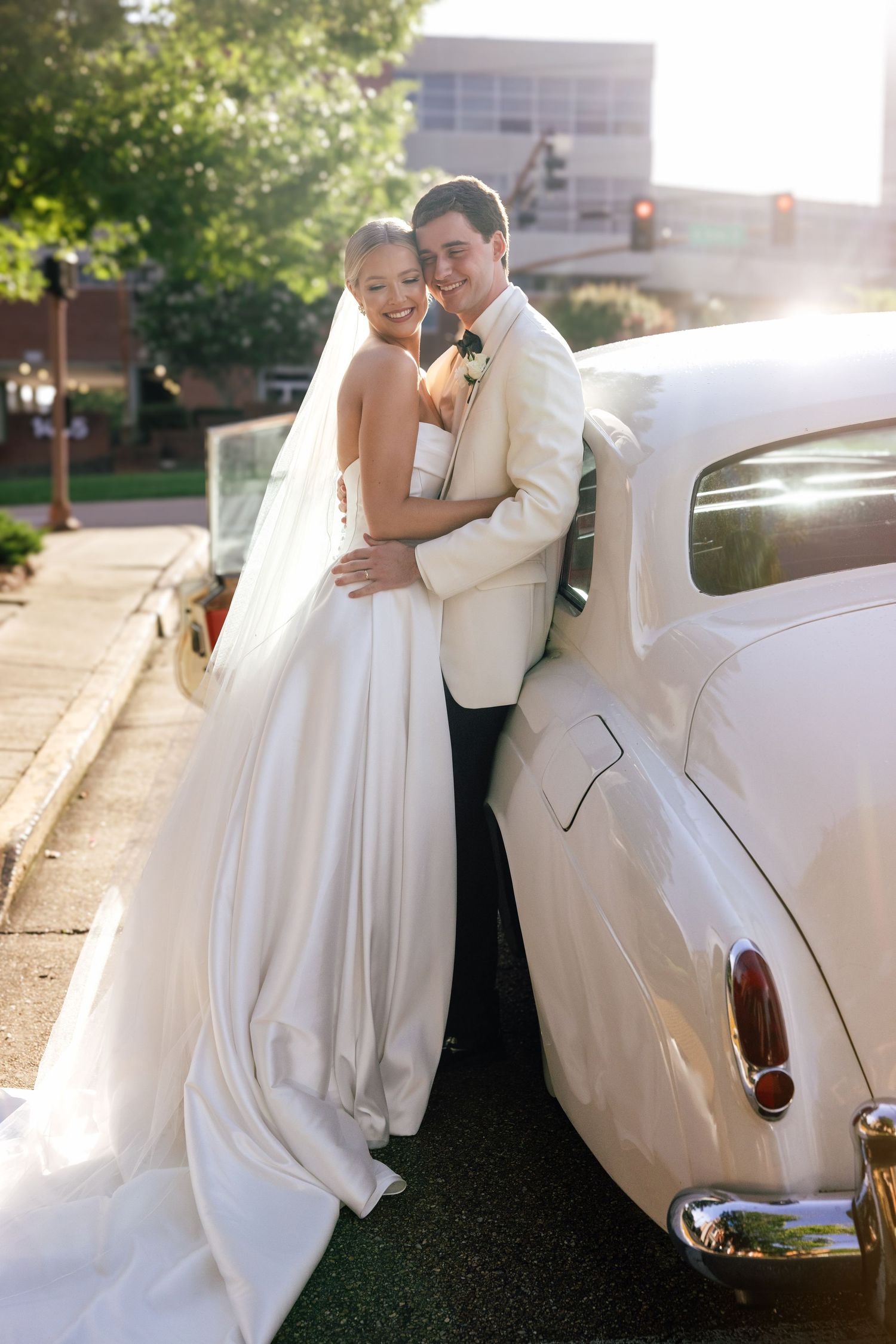 A couple in wedding attire embraces next to a classic white vintage car during sunset.
