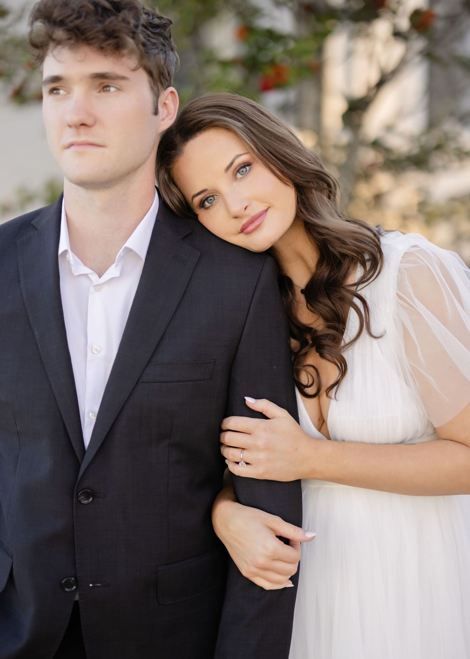 A couple in wedding attire pose together outdoors, with the bride in white leaning against her partner in a black suit.