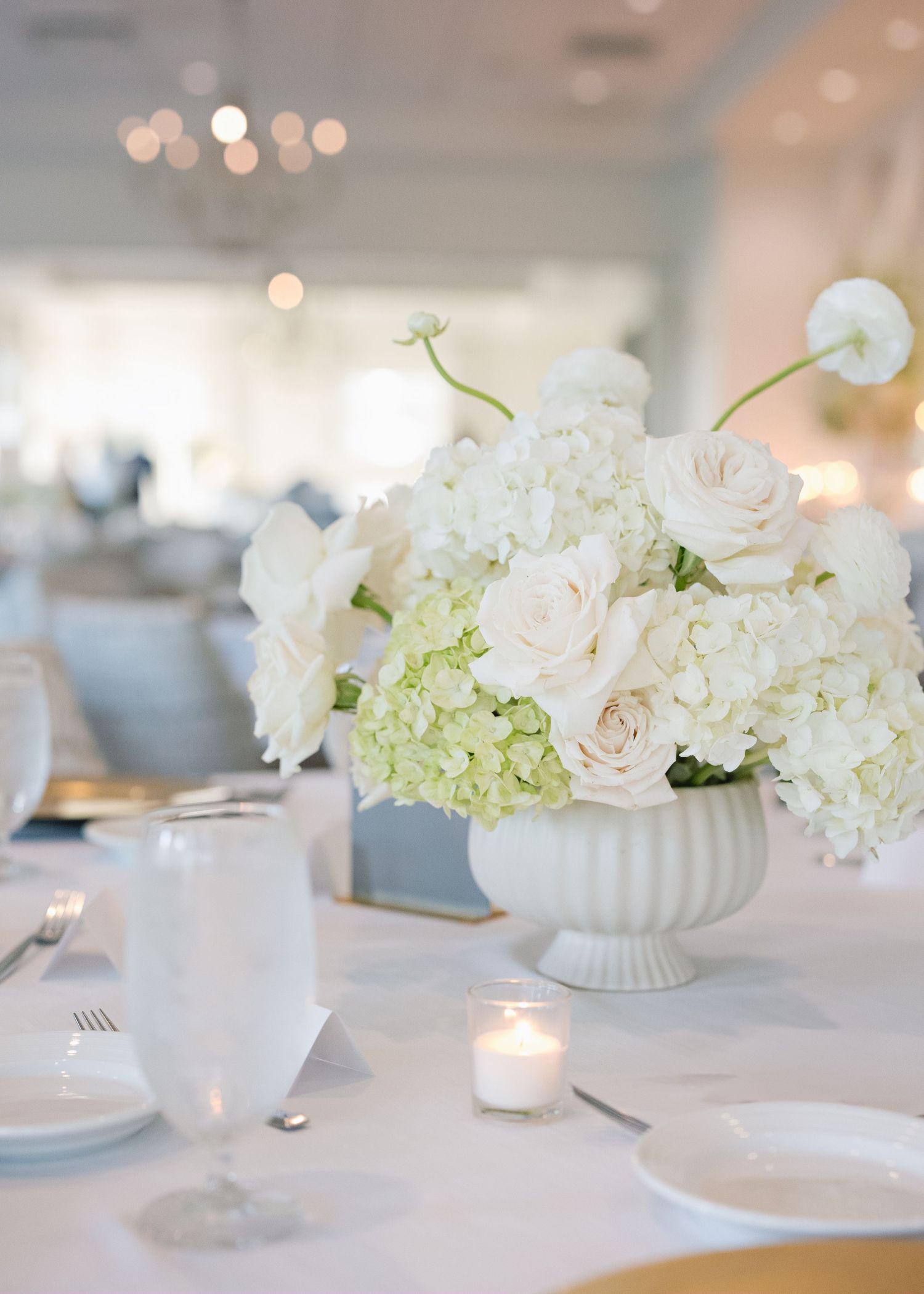 White floral centerpieces in milk glass vases create an elegant table setting with soft candlelight and classic place settings.