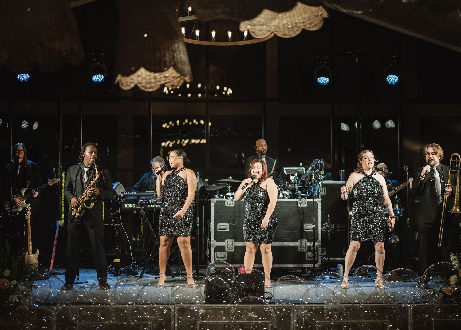 A live band performs on stage with vocalists in sparkly dresses and musicians with instruments against a dark backdrop.