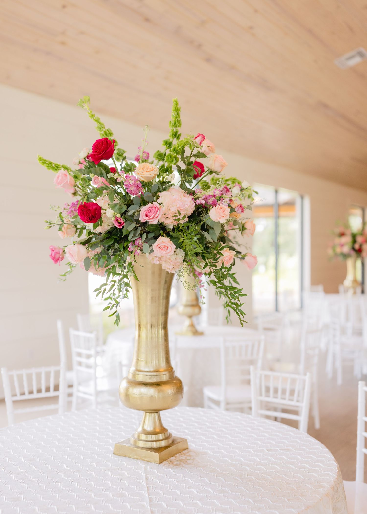 Wedding reception decor features gold vases with pink and red floral arrangements on white tables.